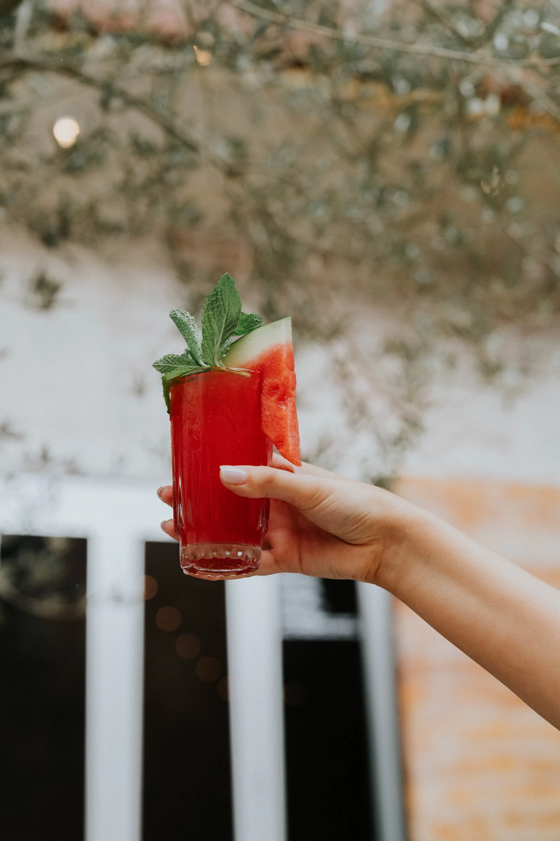A person holding a glass of watermelon cocktail, garnished with a slice of watermelon and a sprig of mint.