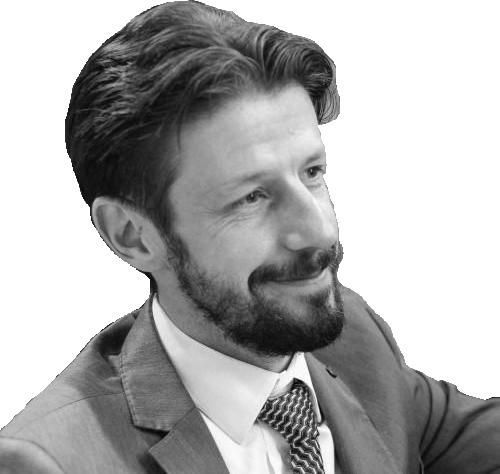 Homme souriant, barbu et moustachu, vêtu d'un costume et d'une cravate à motifs, regardant sur le côté.