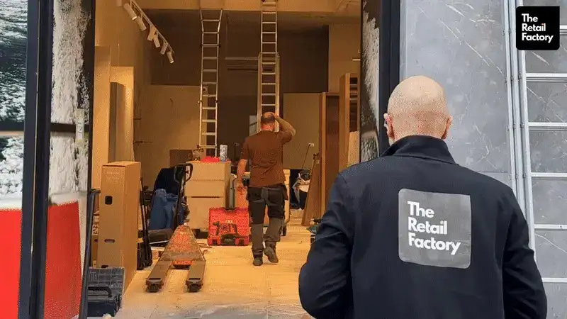 Deux hommes dans un entrepôt ou un grand espace de travail intérieur avec des échelles, des boîtes et du matériel ; l'un d'eux, tournant le dos, porte une veste avec le logo «The Retail Factory