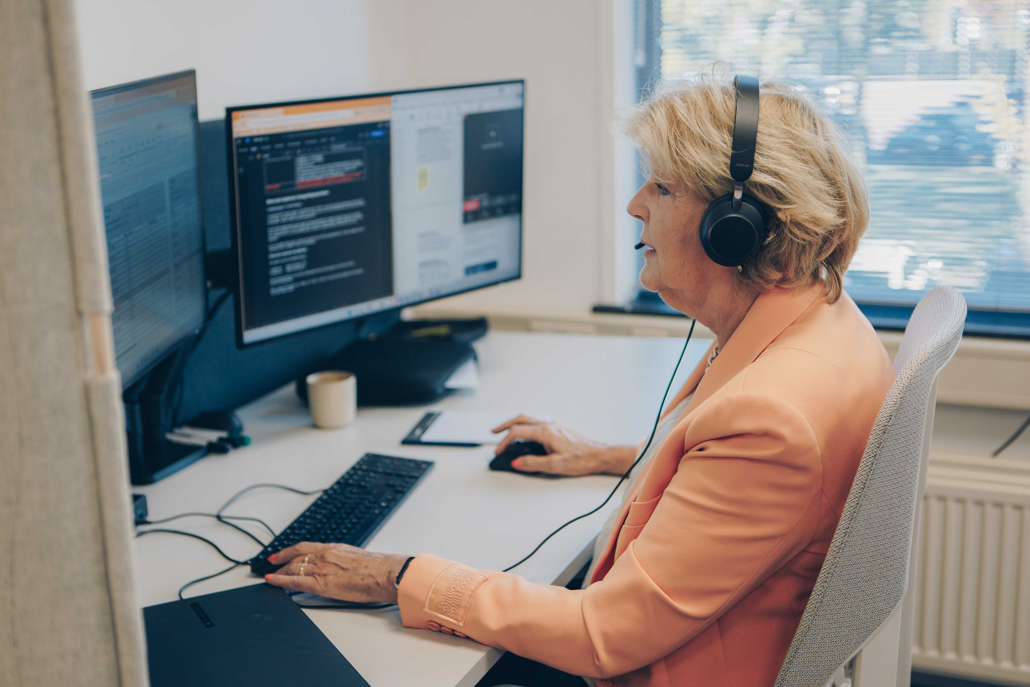 Vrouw met koptelefoon aan het werk achter een bureau met twee computerschermen.