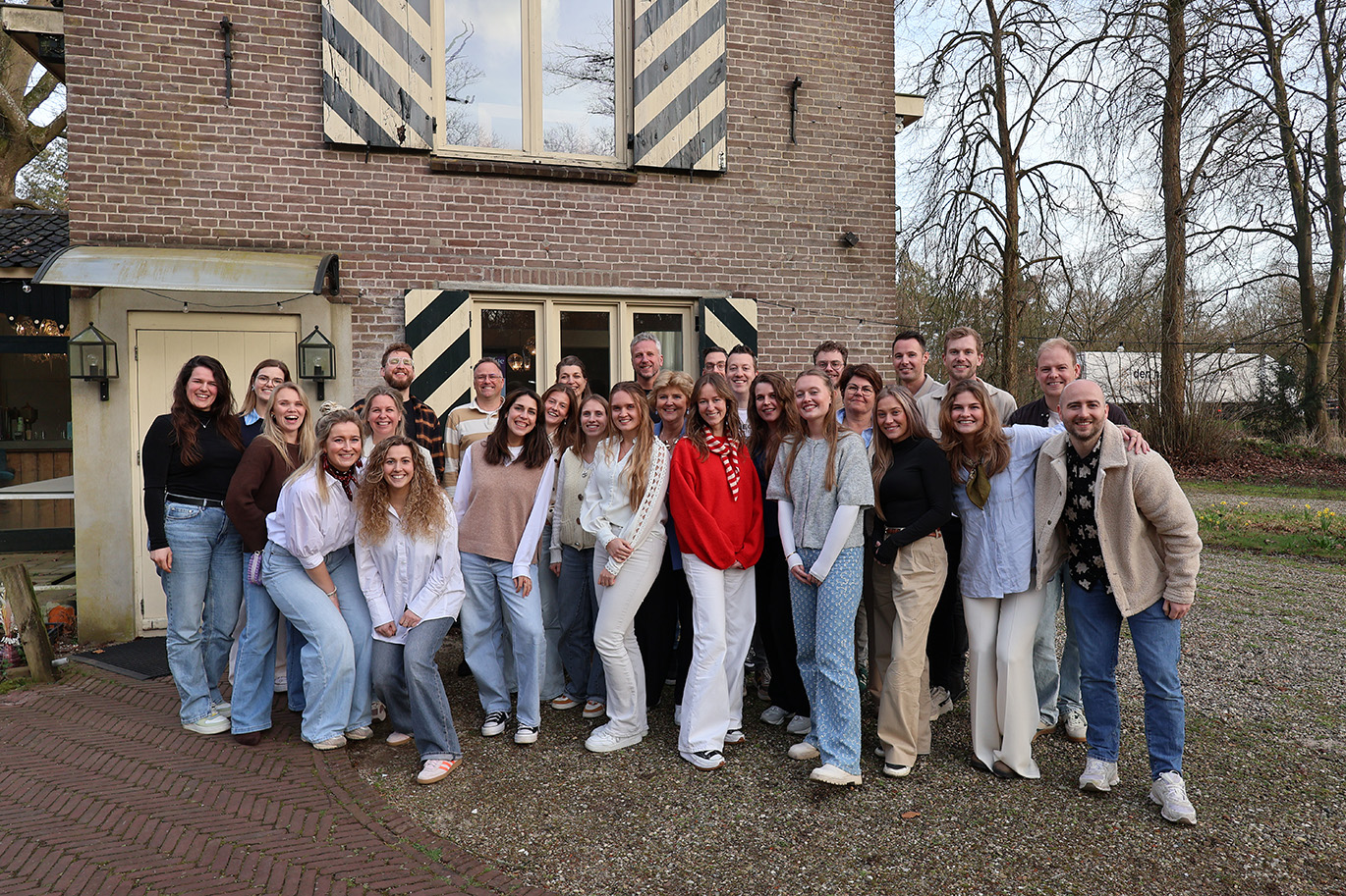 Groep van 27 mensen poseert buiten voor een bakstenen gebouw met ramen.