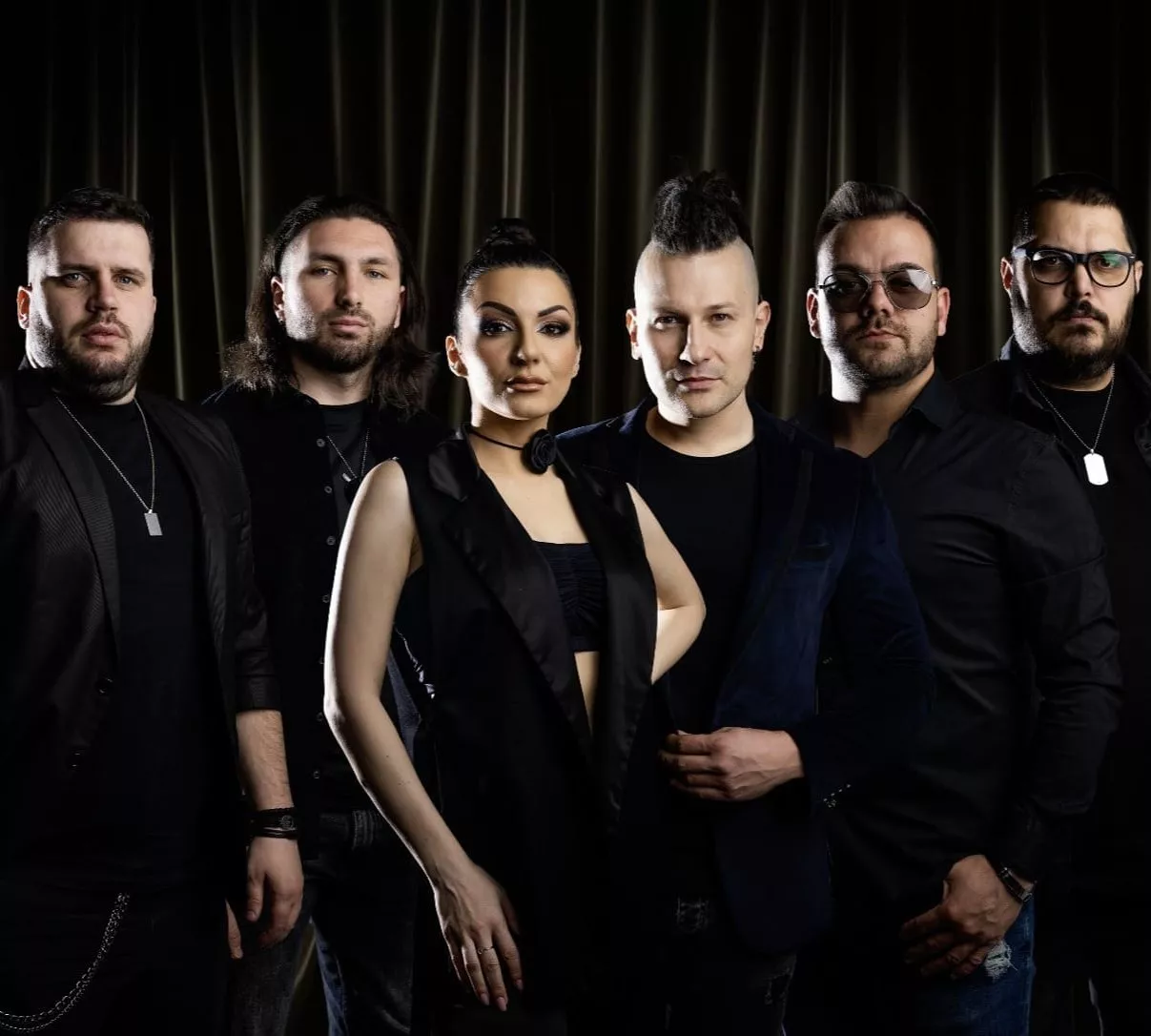 Six people standing in a row against a dark curtain background, five men and one woman, all dressed in black outfits.