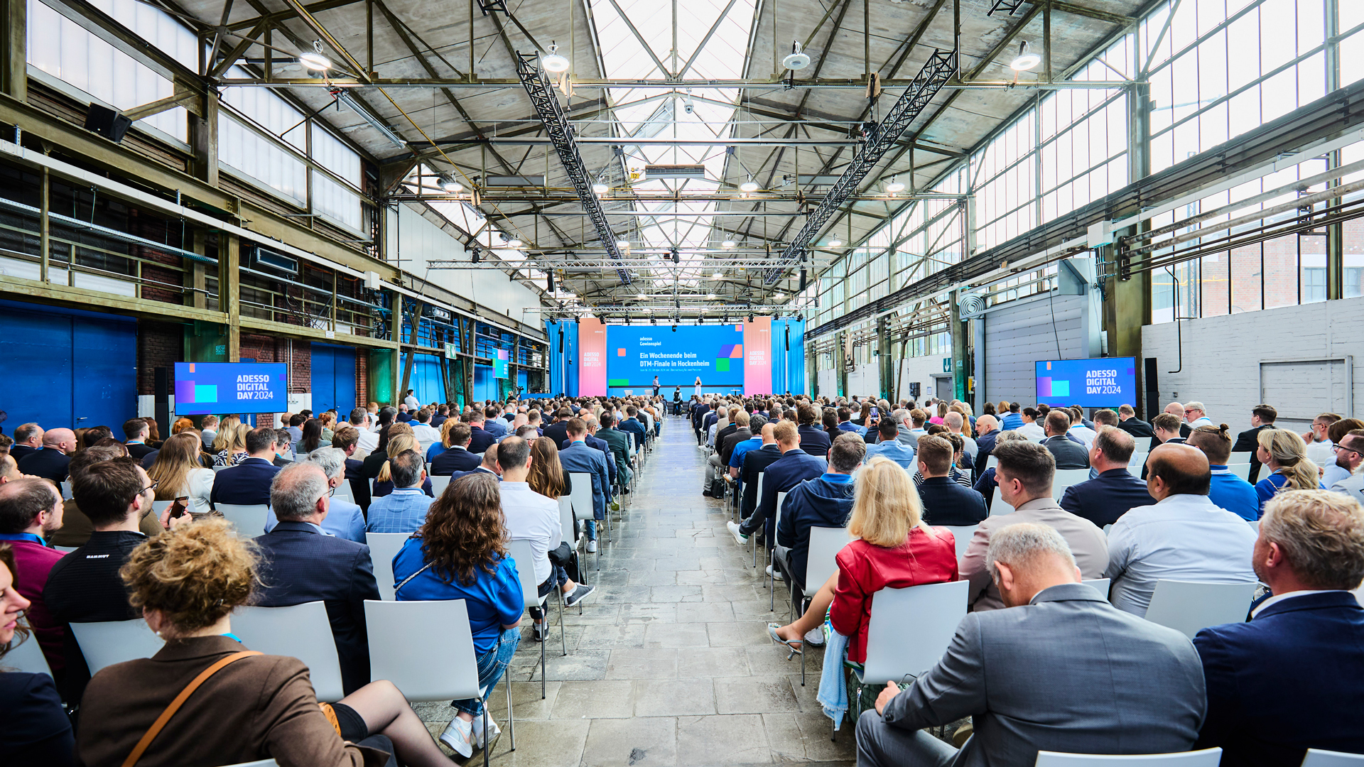Vollbesetzter Konferenzsaal des Adesso Digital Day 2024 in einer Industriehalle: Hunderte Teilnehmende verfolgen die Keynote auf der bunt gestalteten Bühne im Eventdesign von Phocus Brand Contact.