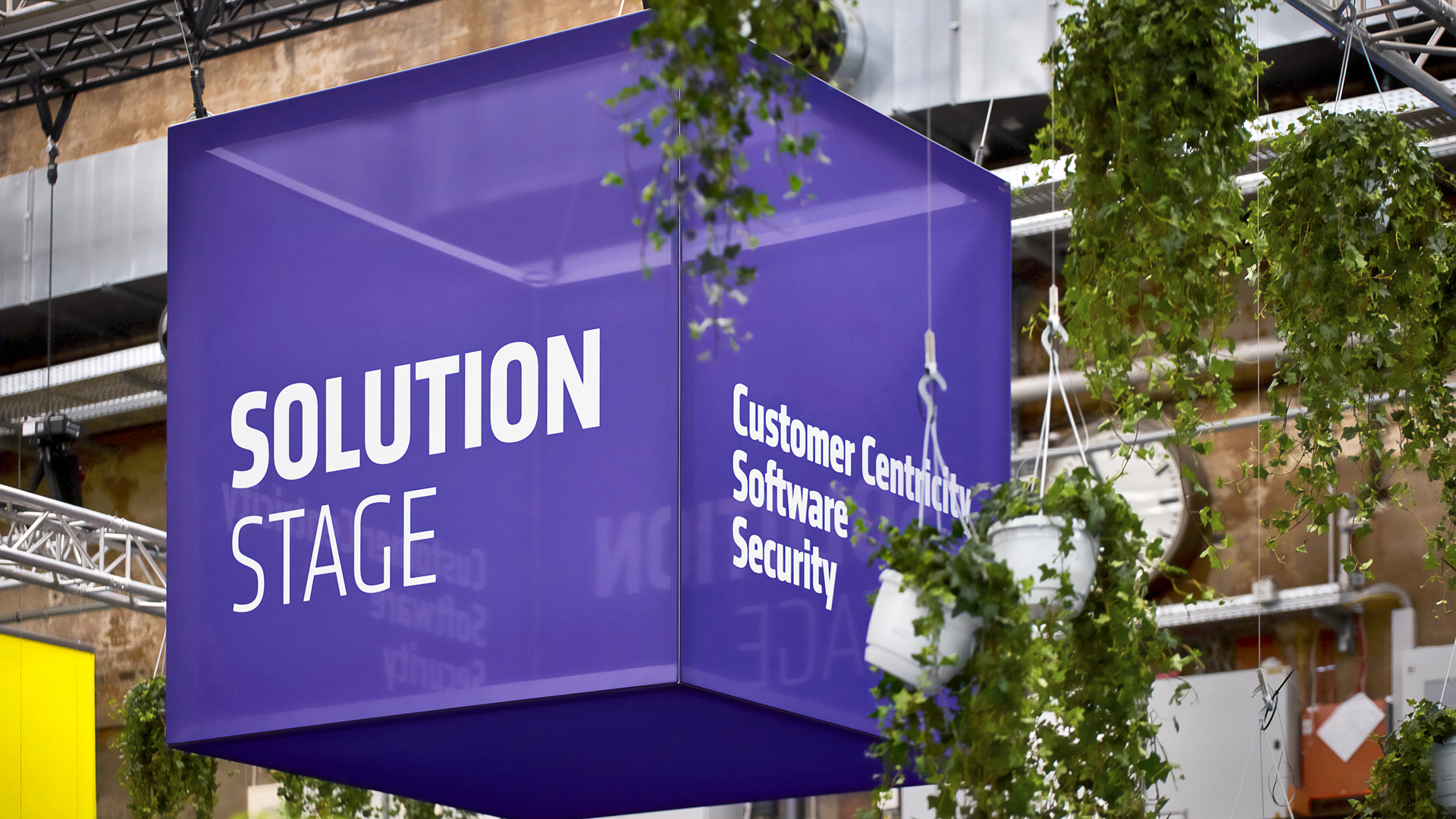 Violetter Hängekubus „Solution Stage" mit den Themen Customer Centricity, Software und Security beim Adesso Digital Day – Eventdesign von Phocus Brand Contact in einer begrünten Industriehalle.