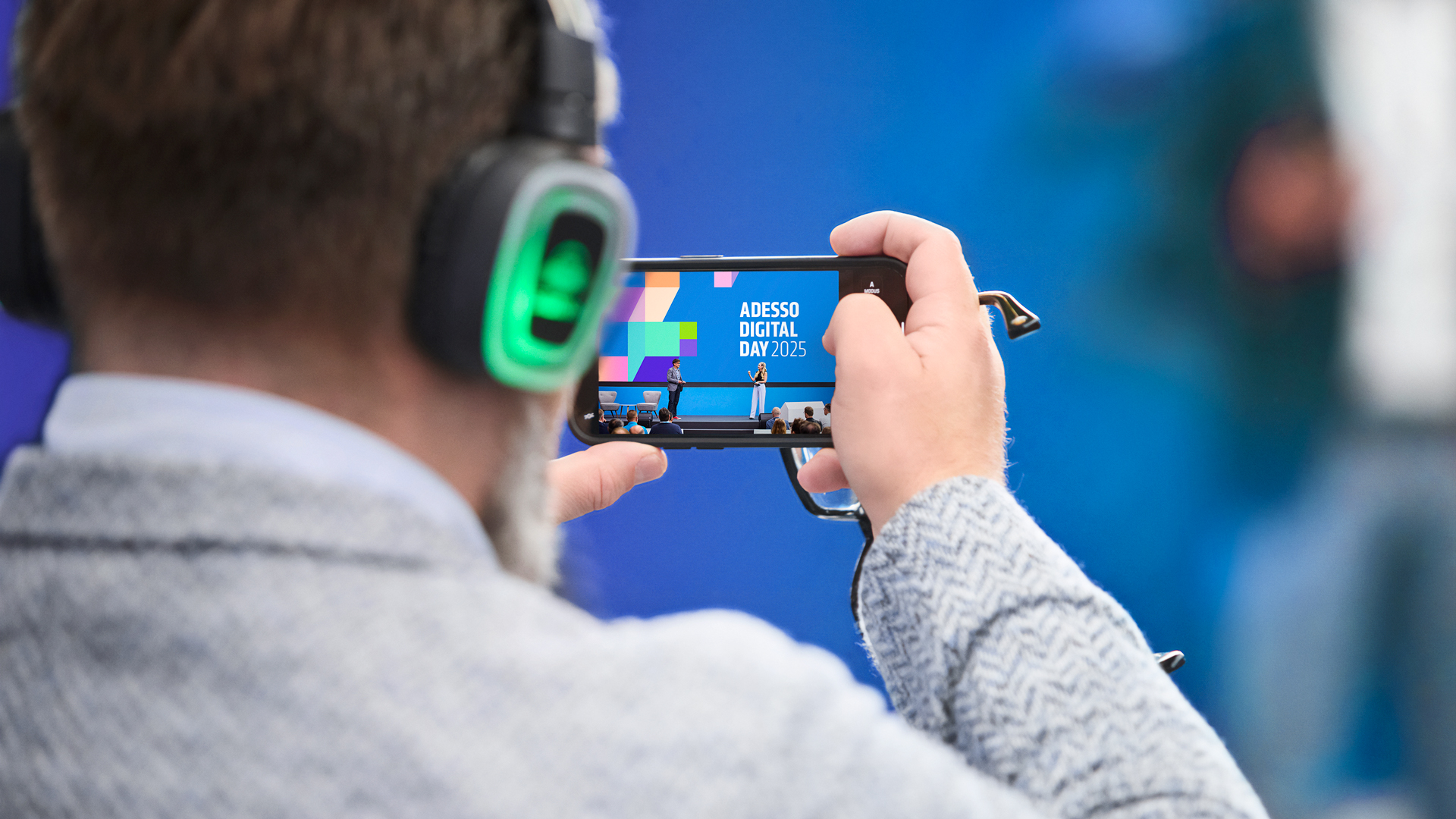Teilnehmer mit Silent-Headset filmt die Bühne des Adesso Digital Day 2025 mit dem Smartphone – im Display das Eventdesign von Phocus Brand Contact mit dem Key-Visual in Blau und Bunt.