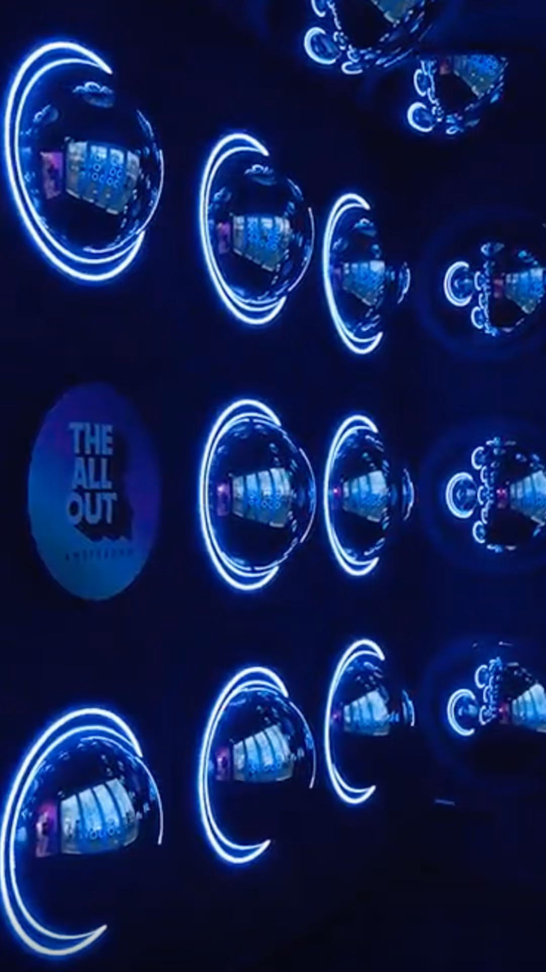 Een donkere muur met een patroon van bolvormige reflecties omgeven door blauwe neonlichten, en een logo van "The All Out Amsterdam".