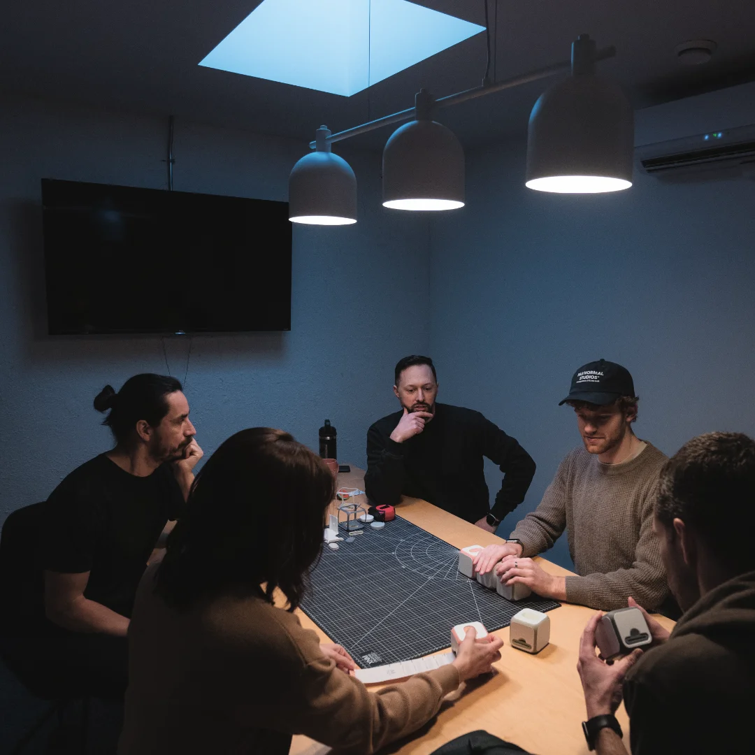 Five people sitting around a table under hanging lamps in a dimly lit room, engaged in a discussion or activity with small cube-shaped objects.