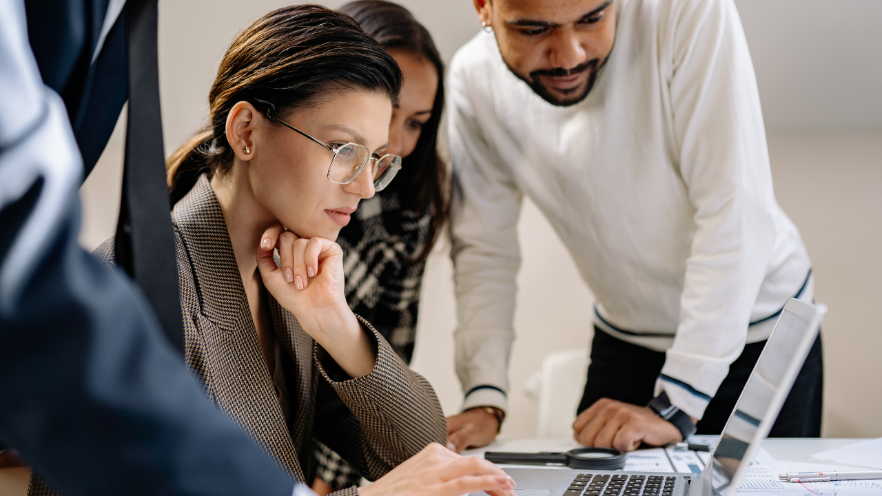 A focused group of three colleagues leans over a white desk, looking intently at a laptop screen. A woman in a grey blazer and glasses sits in the center with her hand on her chin, while two others stand beside her, observing the work among scattered paper