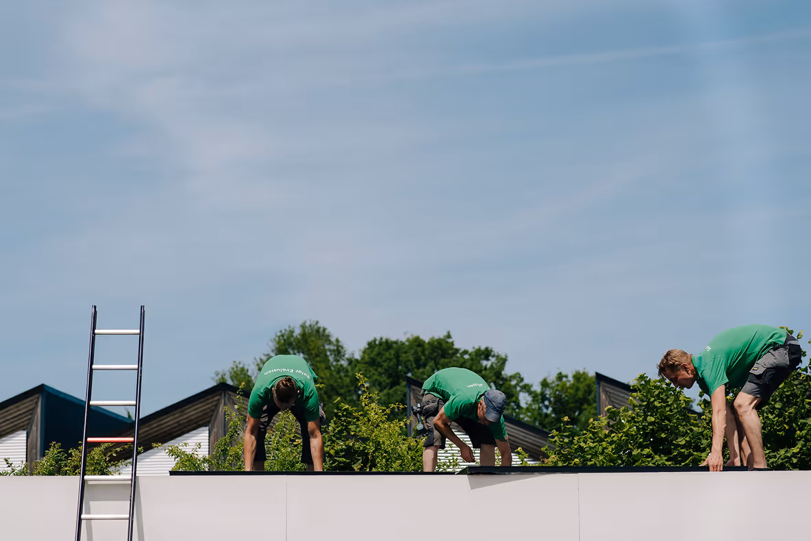Drie installateurs in groene shirts verwijderen zonnepanelen van een plat dak tijdens zonnig weer, met een ladder tegen de gevel en bomen op de achtergrond.