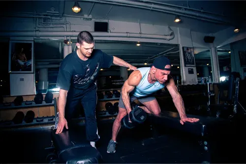 Trainer assists man lifting a 60-pound dumbbell in a dimly lit gym with mirrors and weights.