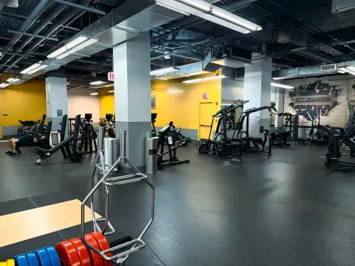 Modern gym with weight plates, machines, benches, and a person lifting weights on the left side.