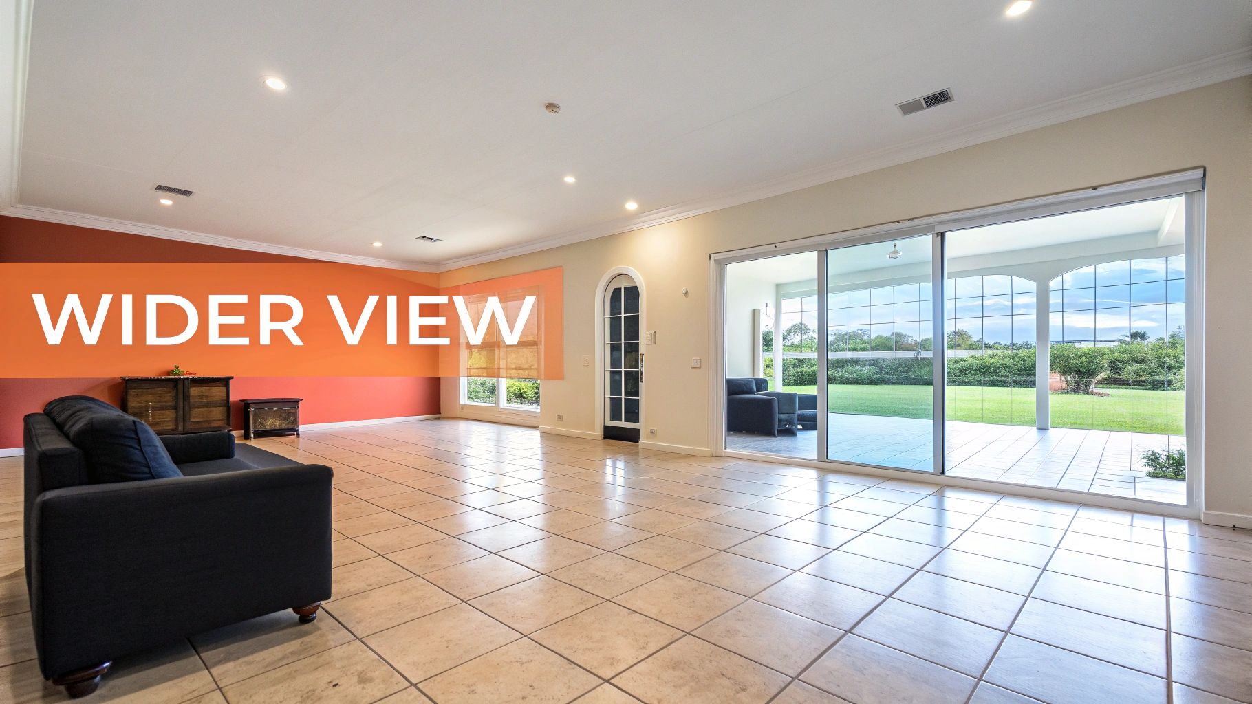 A wide view of a modern living room featuring tile flooring, a dark sofa, and sliding glass doors to a vibrant backyard.