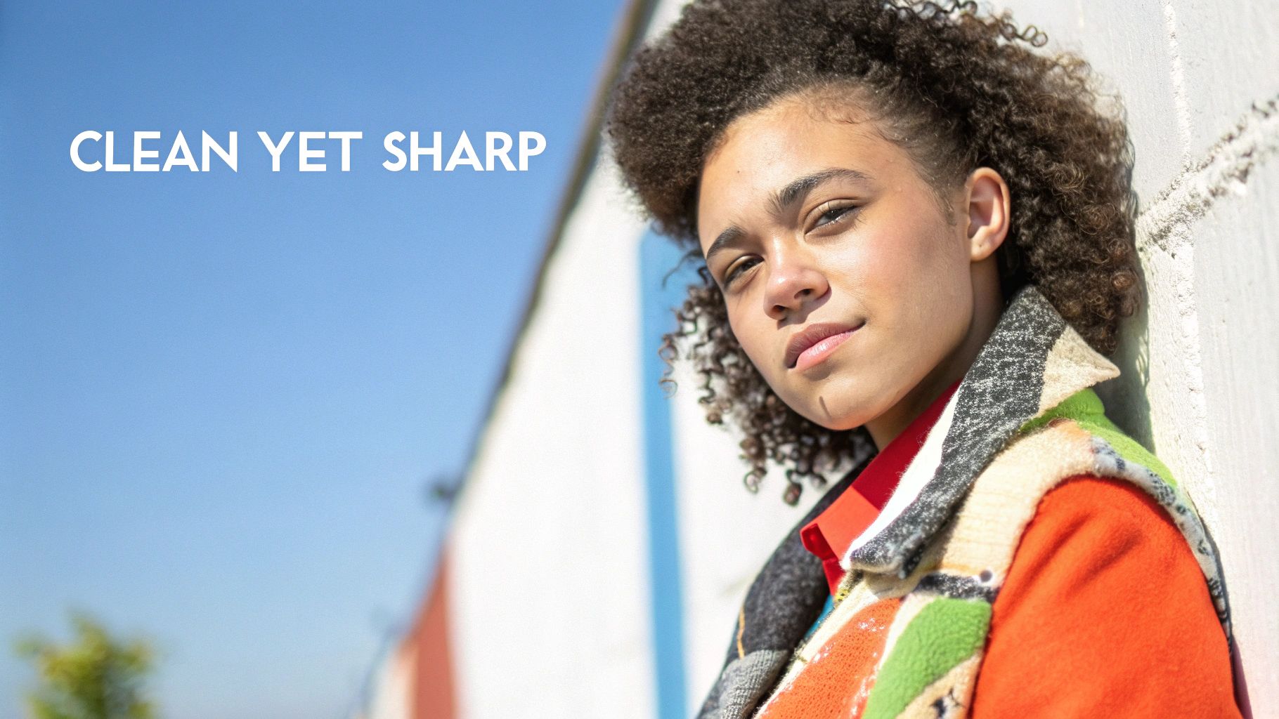 A young woman with curly hair wearing a colorful patterned jacket, leaning against a wall.