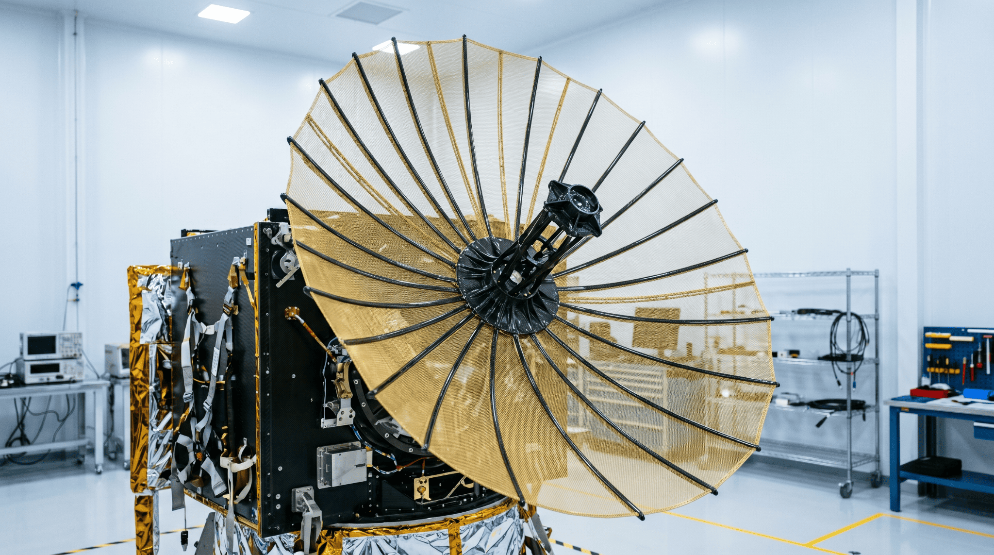 A deployed mesh reflector antenna, its gold reflective mesh stretched over slender carbon-fiber composite ribs that fan from a central hub.