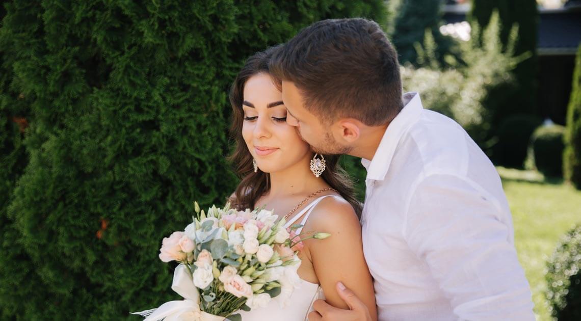Brud ser ned på blomsterbukett mens brudgommen gir henne et kyss på kinnet