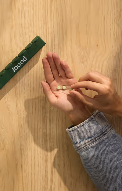 Person wearing a denim jacket holding small green pills in one hand and picking one with the other over a wooden surface, with a green pill organizer labeled 'found' nearby.