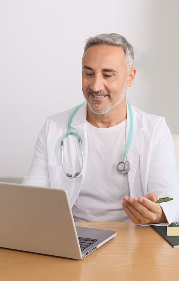 Smiling male doctor with gray hair and a beard wearing a white coat and stethoscope, working on a laptop at a wooden desk.