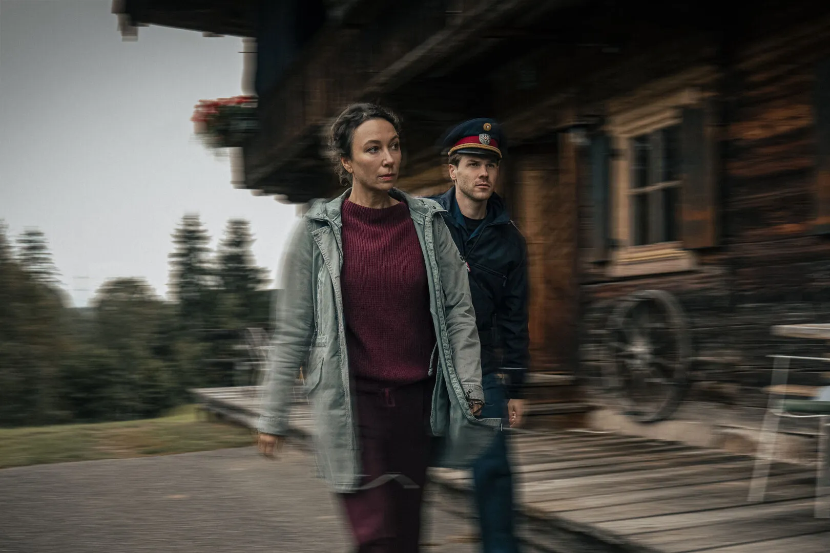 A woman in a light green jacket and burgundy sweater walks with purpose in the foreground, while a uniformed police officer follows closely behind her. Both look focused and serious. They move past a rustic wooden house, with motion blur emphasizing their 