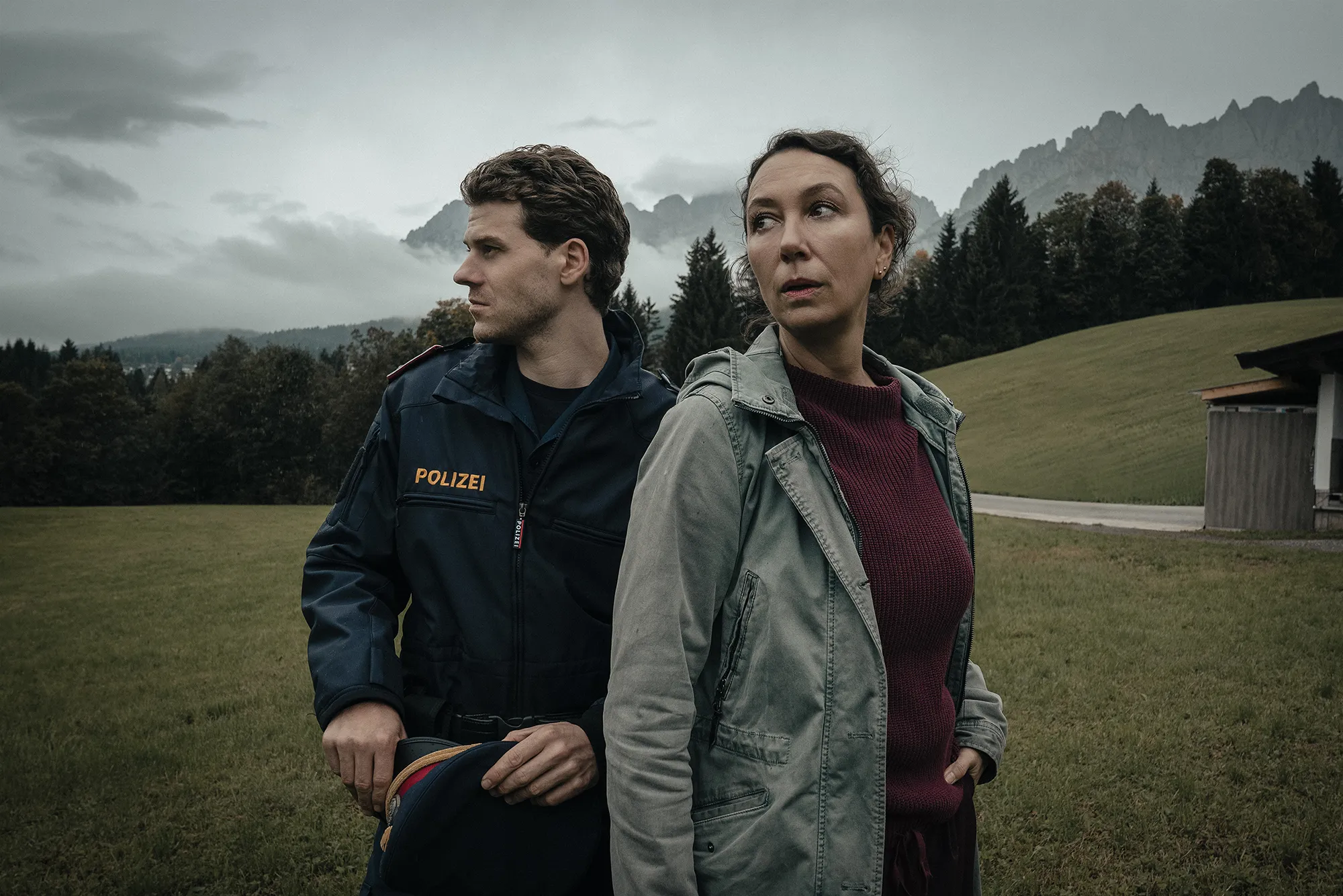 Two people stand side by side in a misty rural landscape, both looking in opposite directions with tense, alert expressions. The man on the left wears a dark police uniform with “Polizei” on the chest, while the woman on the right wears a green jacket over