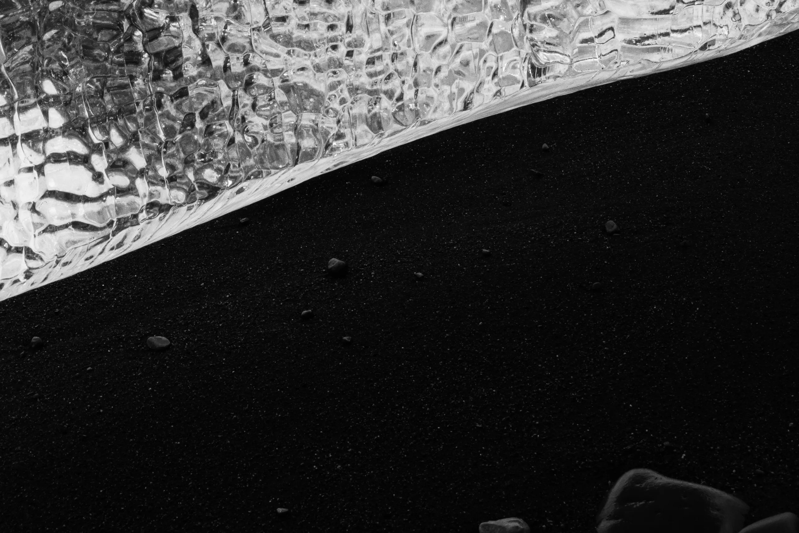 Close-up black and white photograph of a large, intricately textured piece of clear glacier ice resting on fine volcanic black sand. The ice displays a complex, crystalline pattern with ripples and refracted light, creating a high-contrast visual against the dark granular surface.