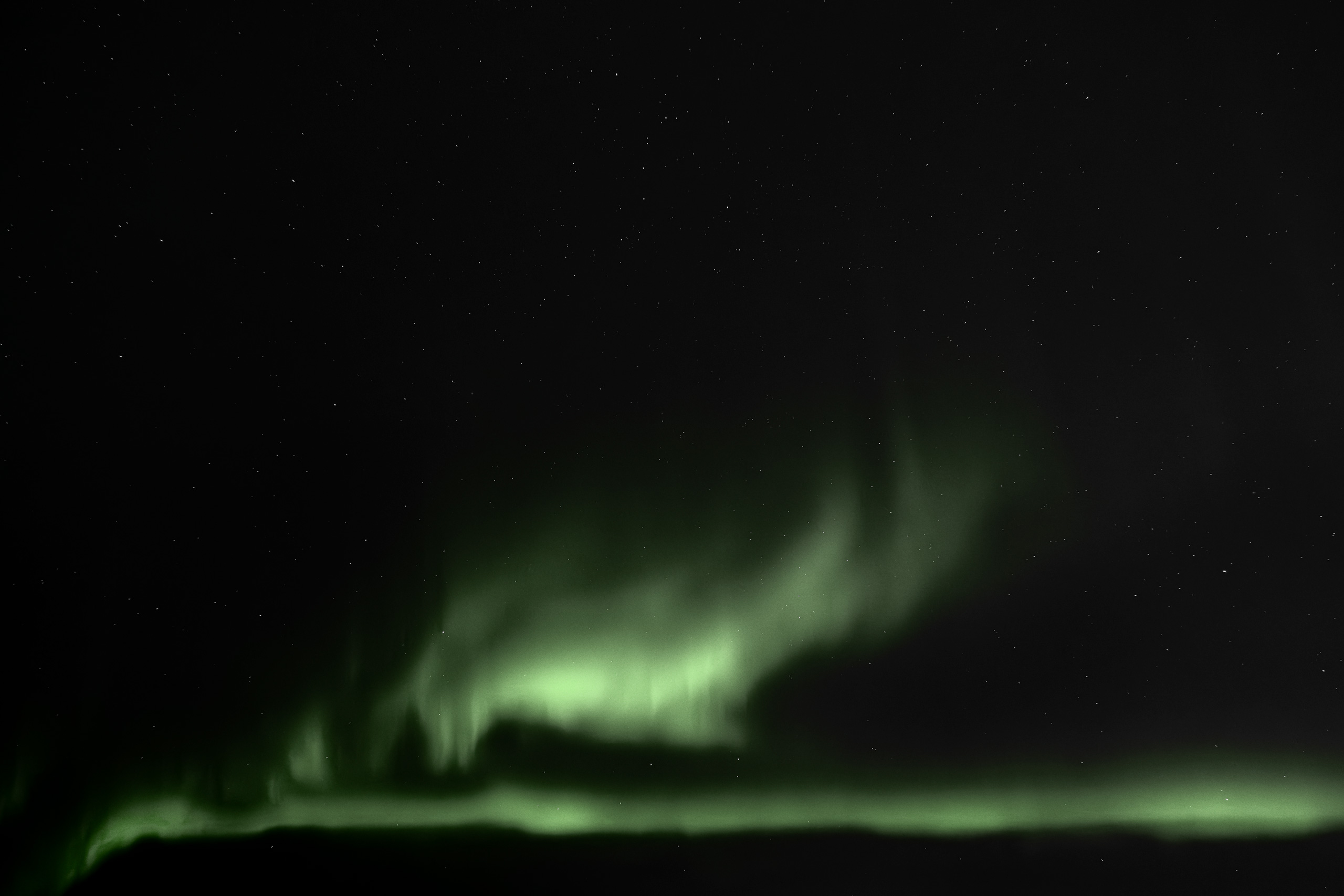 Stunning nighttime photo of the Northern Lights (Aurora Borealis) stretching across a star-dotted sky, with vivid green aurora curtains and subtle vertical rays illuminating the horizon. The lower edge of the frame features a dark silhouette of the landscape, enhancing the contrast of the celestial display.