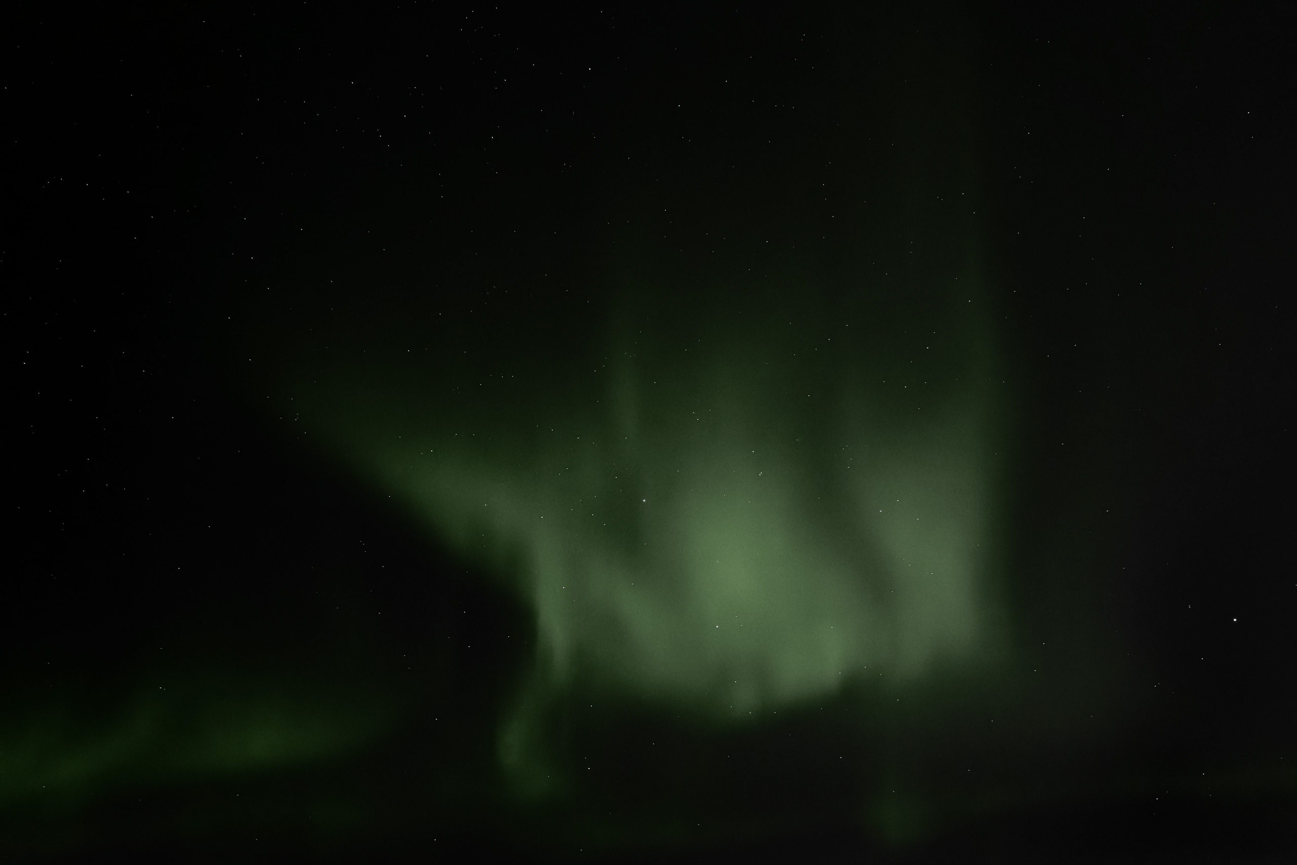 Long-exposure photo of the Northern Lights (Aurora Borealis) illuminating the night sky with ethereal green curtains and rays against a backdrop of visible stars. The aurora arcs upward in vertical streaks, radiating a soft glow and creating a mystical atmosphere.