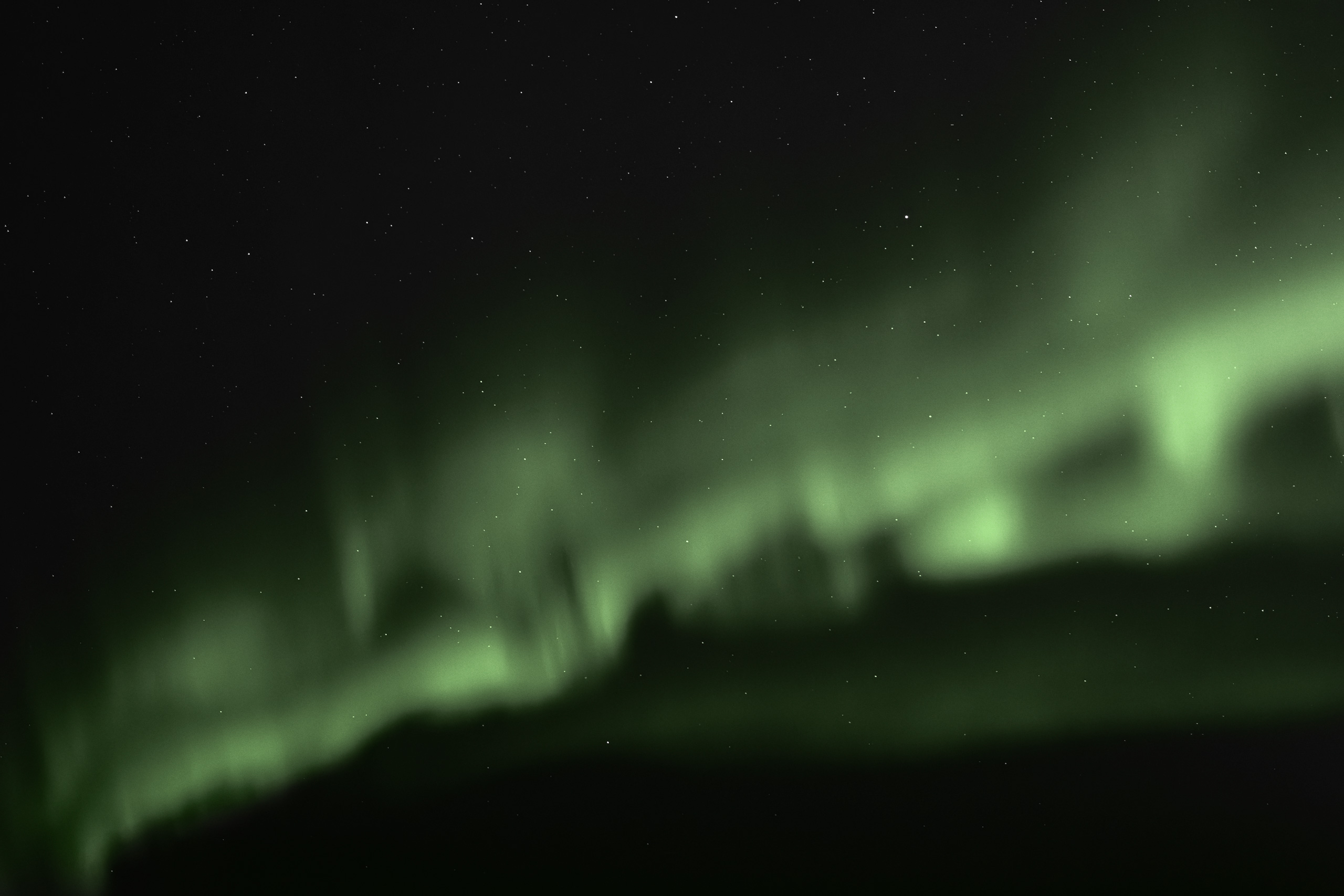 Nighttime photograph of the Northern Lights (Aurora Borealis) illuminating a dark, star-filled sky with swirling green light ribbons. The ethereal glow dances above a faint horizon silhouette, blending atmospheric luminosity with cosmic stillness.