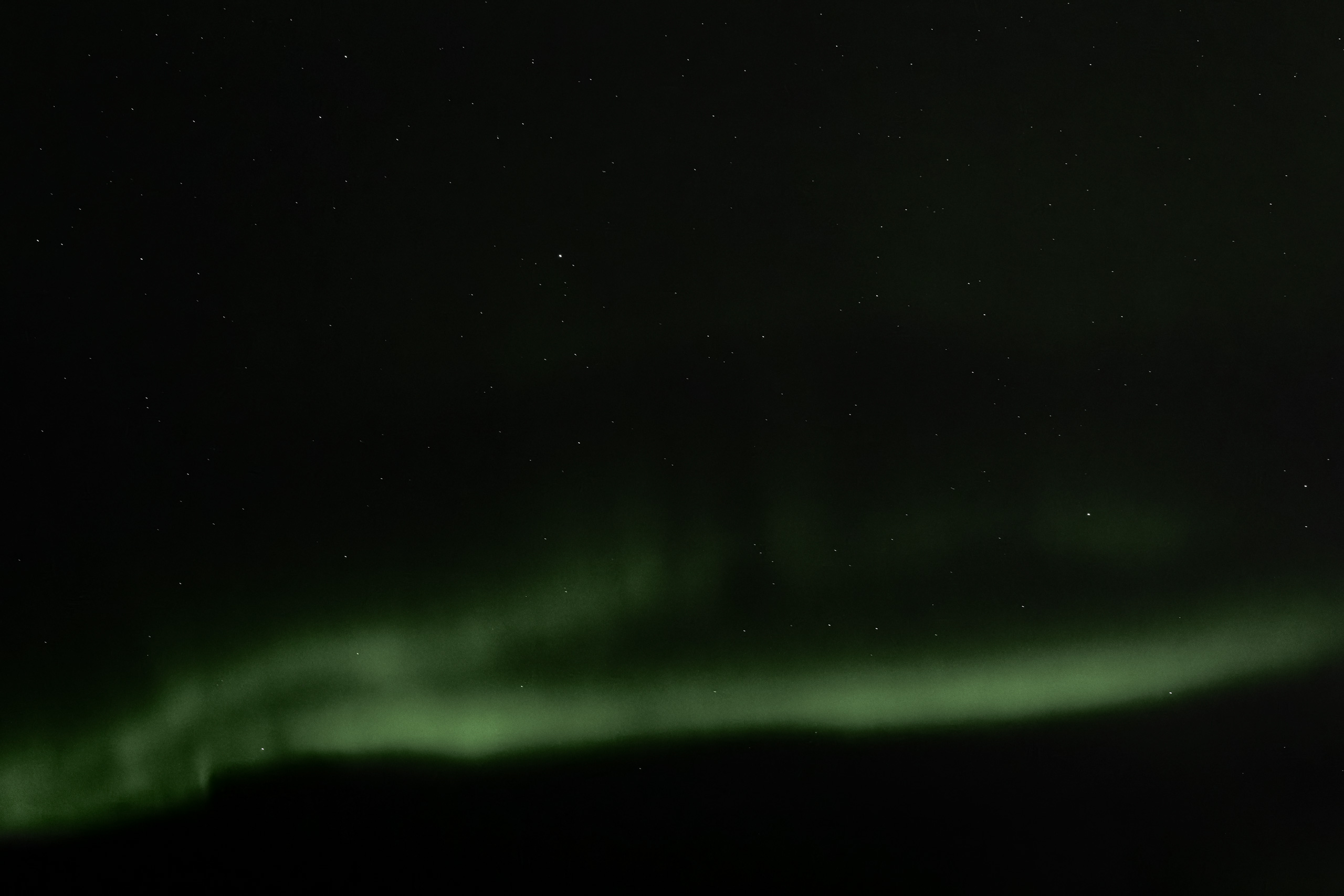 Nighttime photograph capturing a soft green band of the Northern Lights (Aurora Borealis) stretching horizontally across the sky above a dark, silhouetted landscape. The sky is sprinkled with stars, enhancing the tranquil and mystical ambiance.