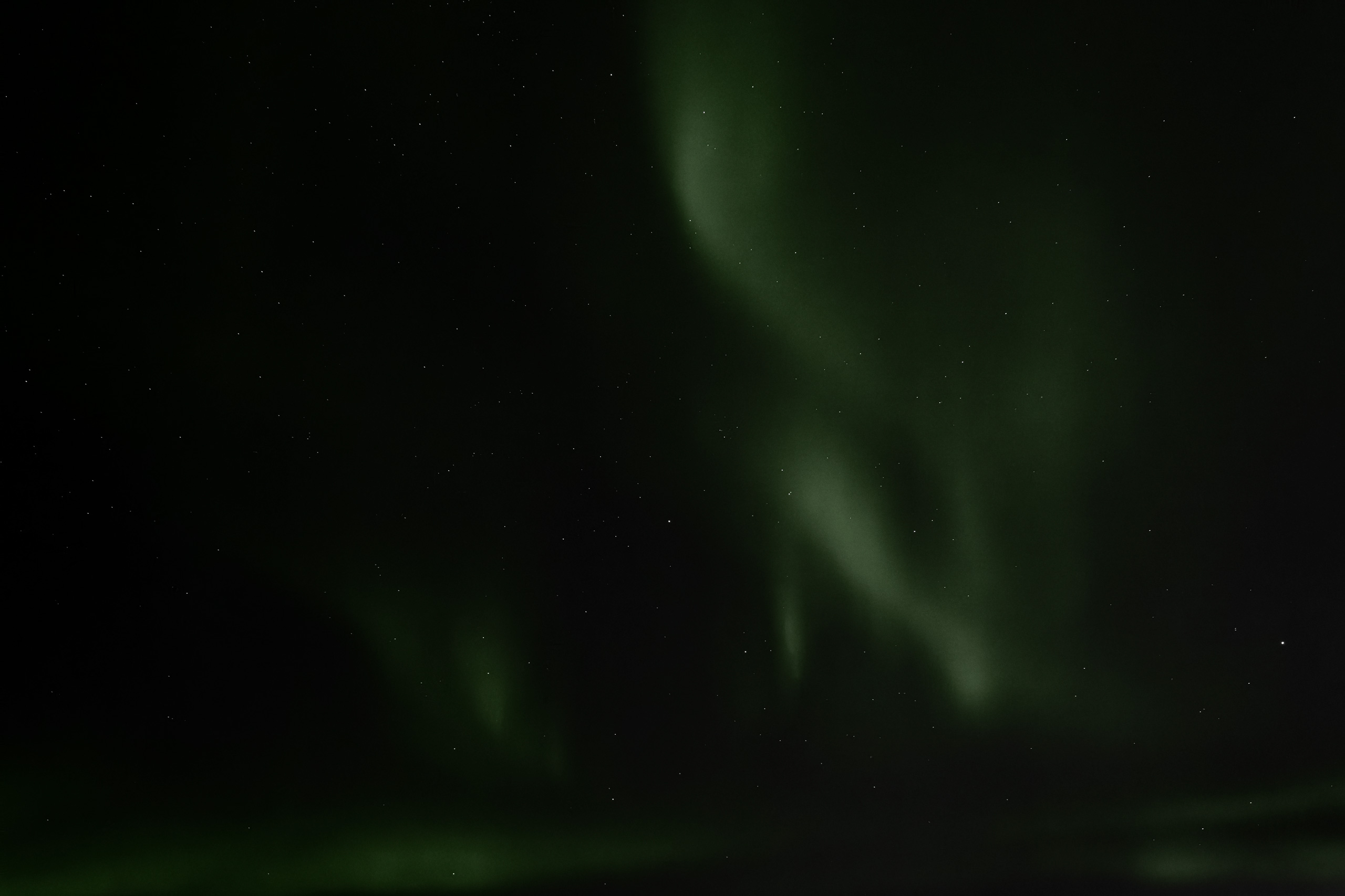 Wide-angle photograph of the Northern Lights displaying soft green aurora streaks and ribbons against a starry night sky. The wisps of aurora arc gently upward, creating a fluid, wave-like formation across the dark celestial backdrop.