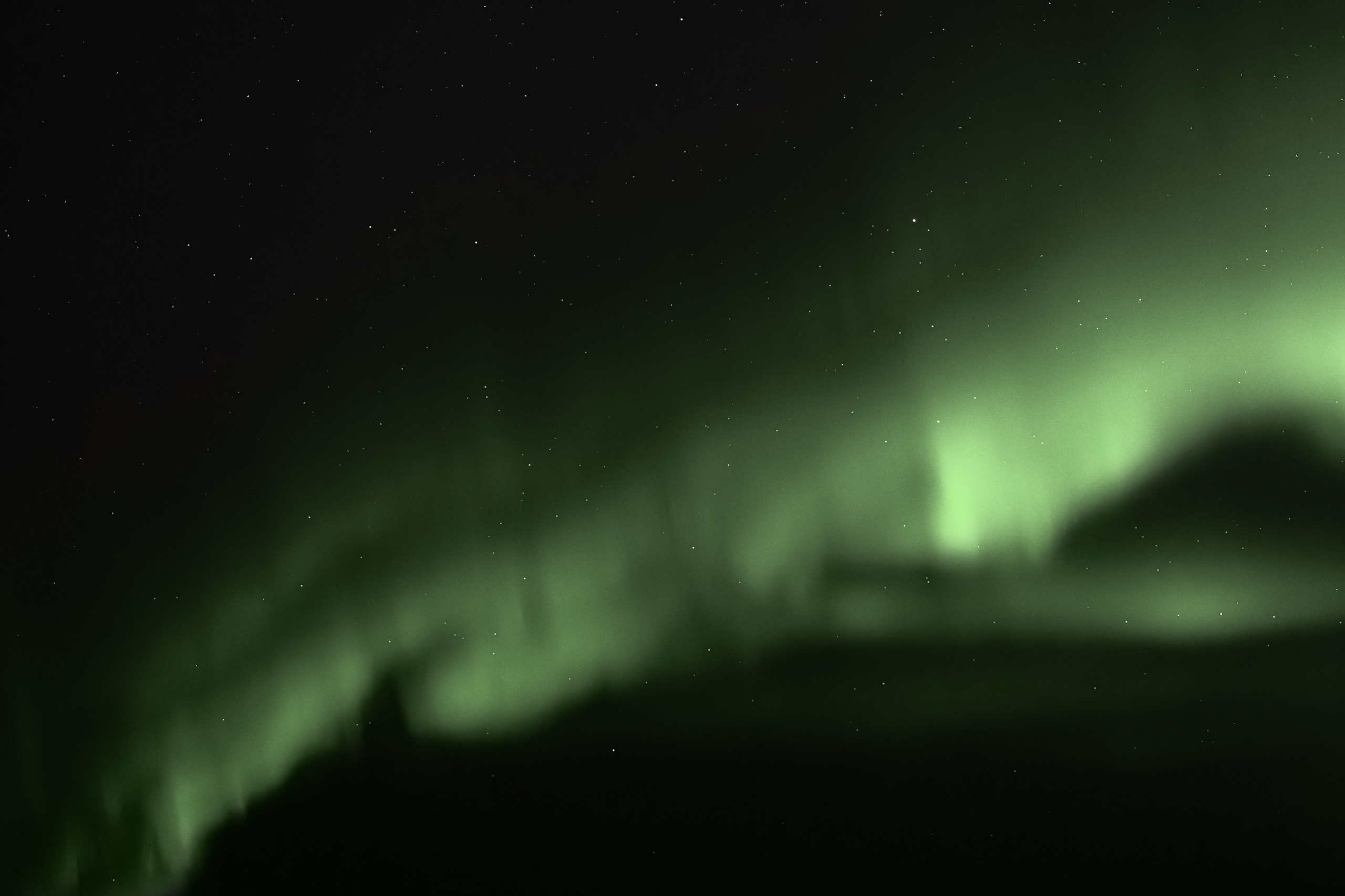 Landscape photograph of the Aurora Borealis, also known as the Northern Lights, glowing in vivid green waves across a starry night sky. The aurora forms fluid, atmospheric curtains of light above a dark, silhouetted horizon, creating a sense of wonder and natural beauty.