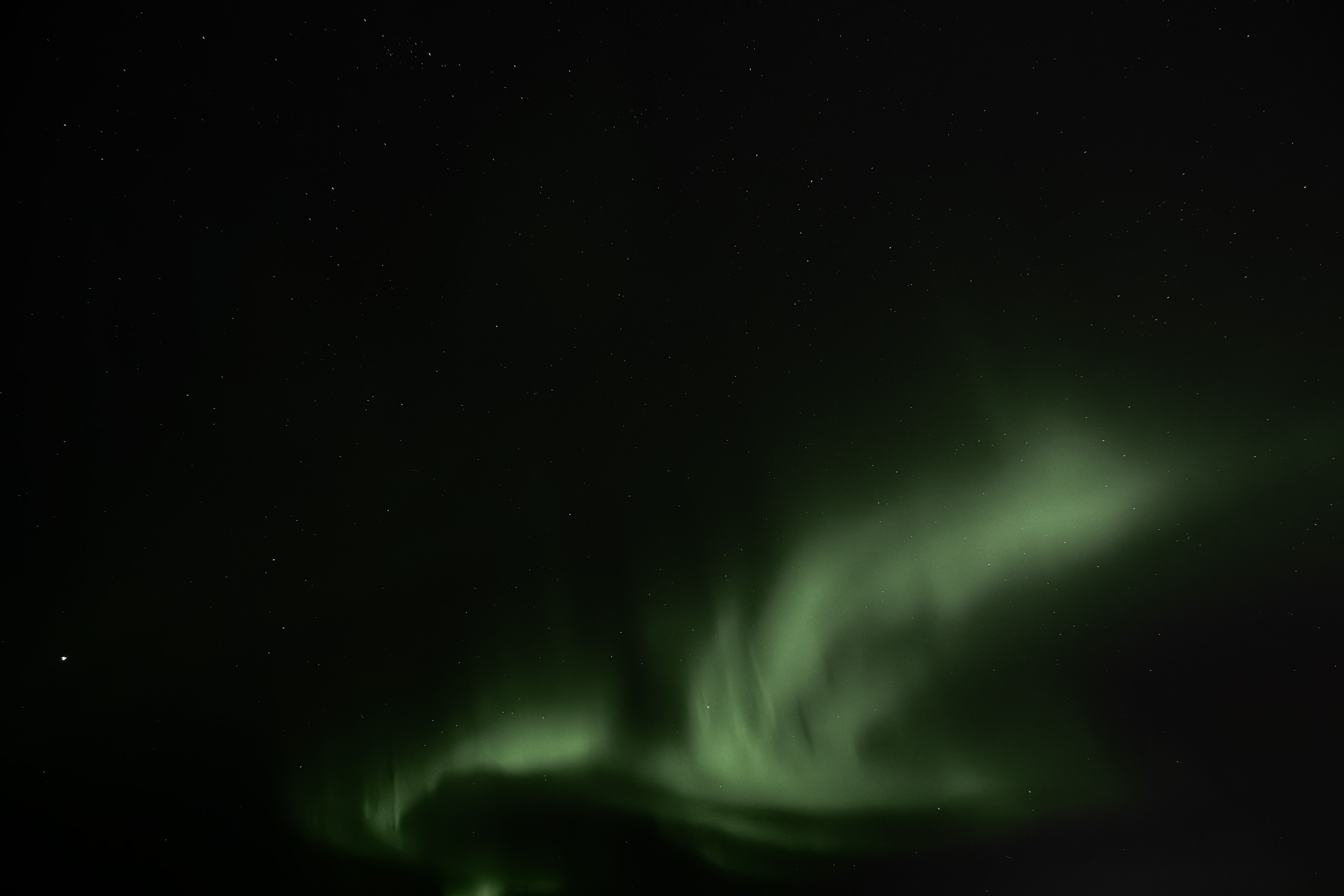 Long-exposure photograph of the Northern Lights showing vivid green aurora streaks swirling and dancing across a star-filled night sky. The luminous bands of light arc and twist in fluid patterns above a dark, silhouetted horizon.