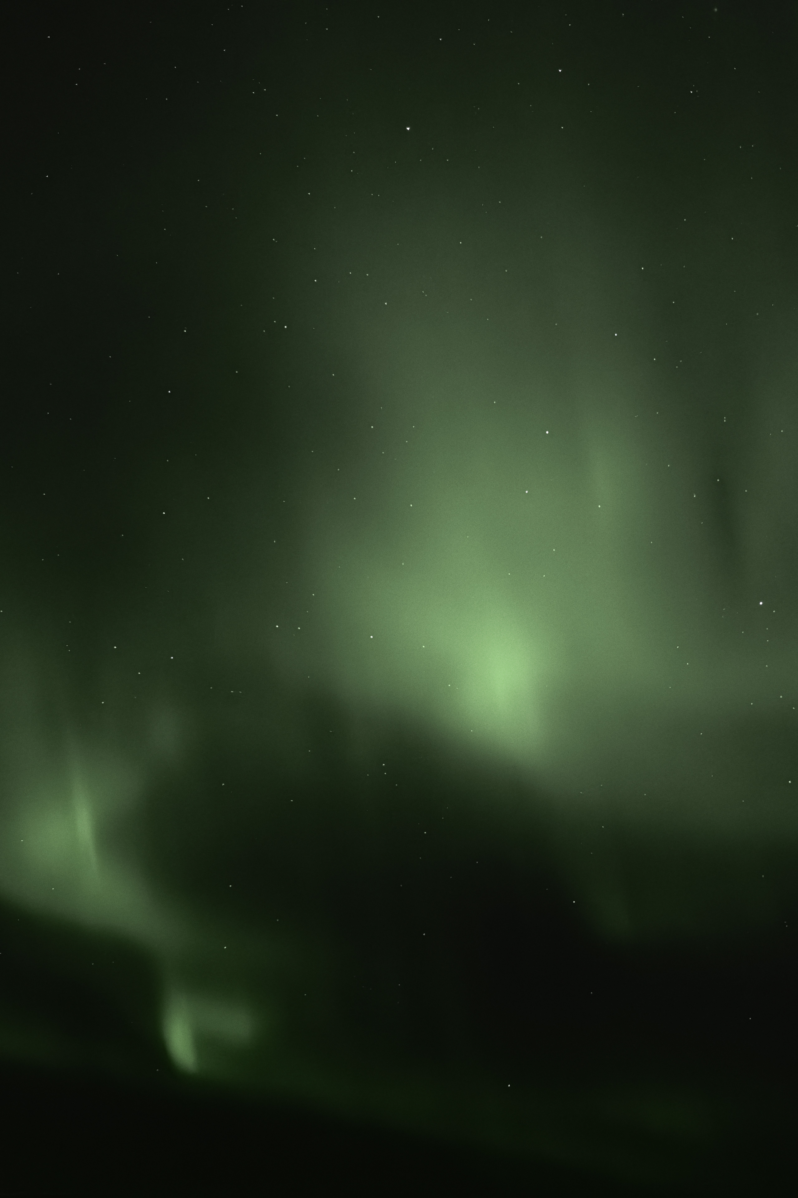 Photograph of the Northern Lights displaying glowing green aurora streaks and wisps across a dark, starry night sky. The aurora borealis forms a soft, ethereal veil of light, subtly illuminating the atmosphere with a natural, celestial glow.