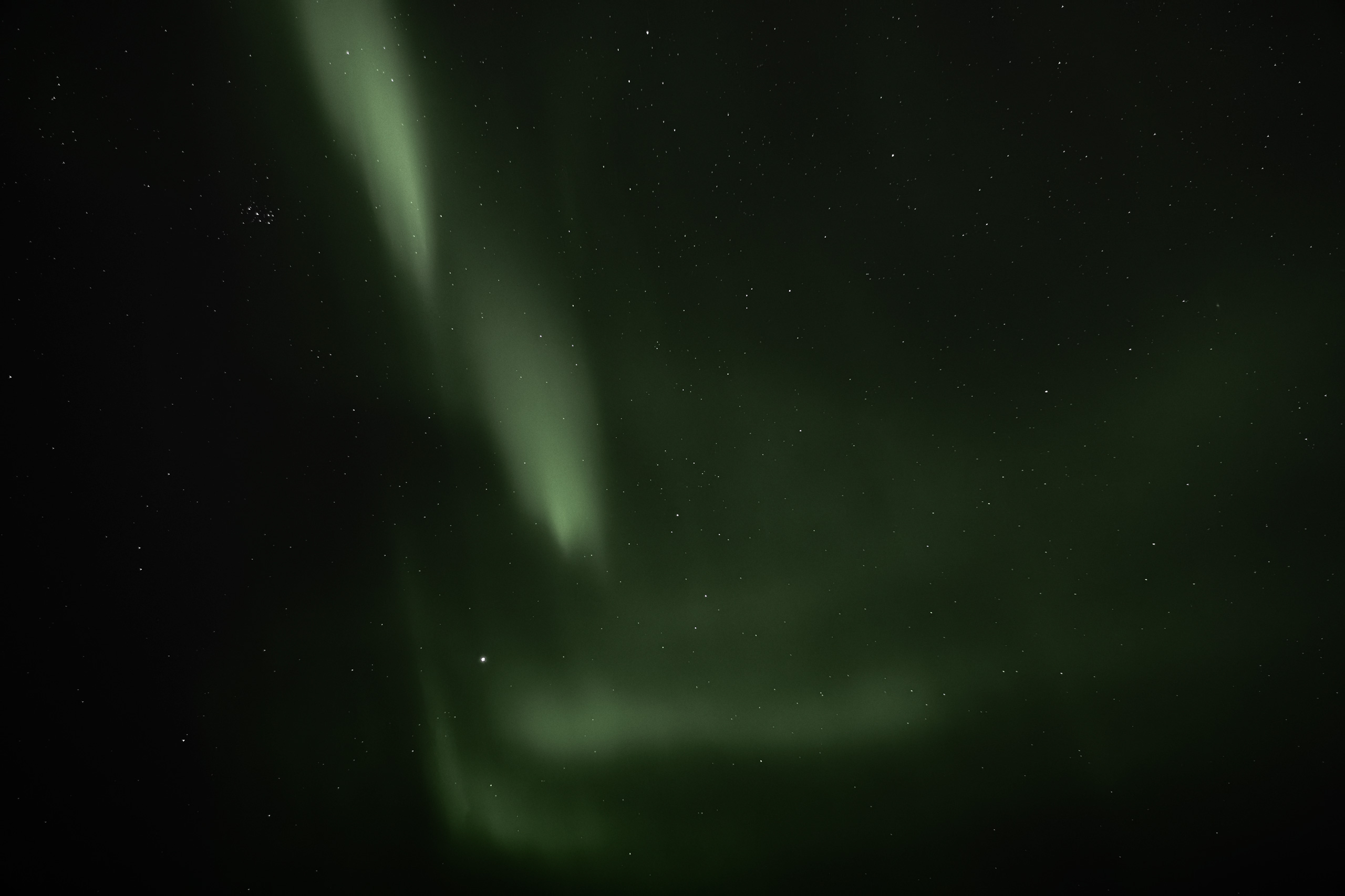 Long-exposure night sky photograph showcasing soft green ribbons of the aurora borealis swirling against a backdrop of countless stars. The Northern Lights appear as faint, glowing arcs moving across the dark Arctic sky.