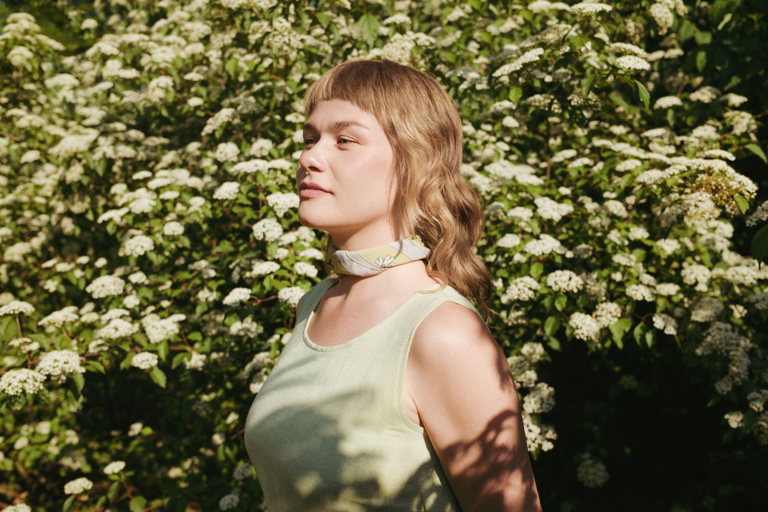 Portrait of a woman standing in front of lush greenery filled with small white blossoms, basking in warm sunlight. She has wavy light brown hair with bangs, wears a pale yellow sleeveless top, and a light floral scarf tied around her neck. The soft lighting and natural setting evoke a serene, springtime atmosphere.