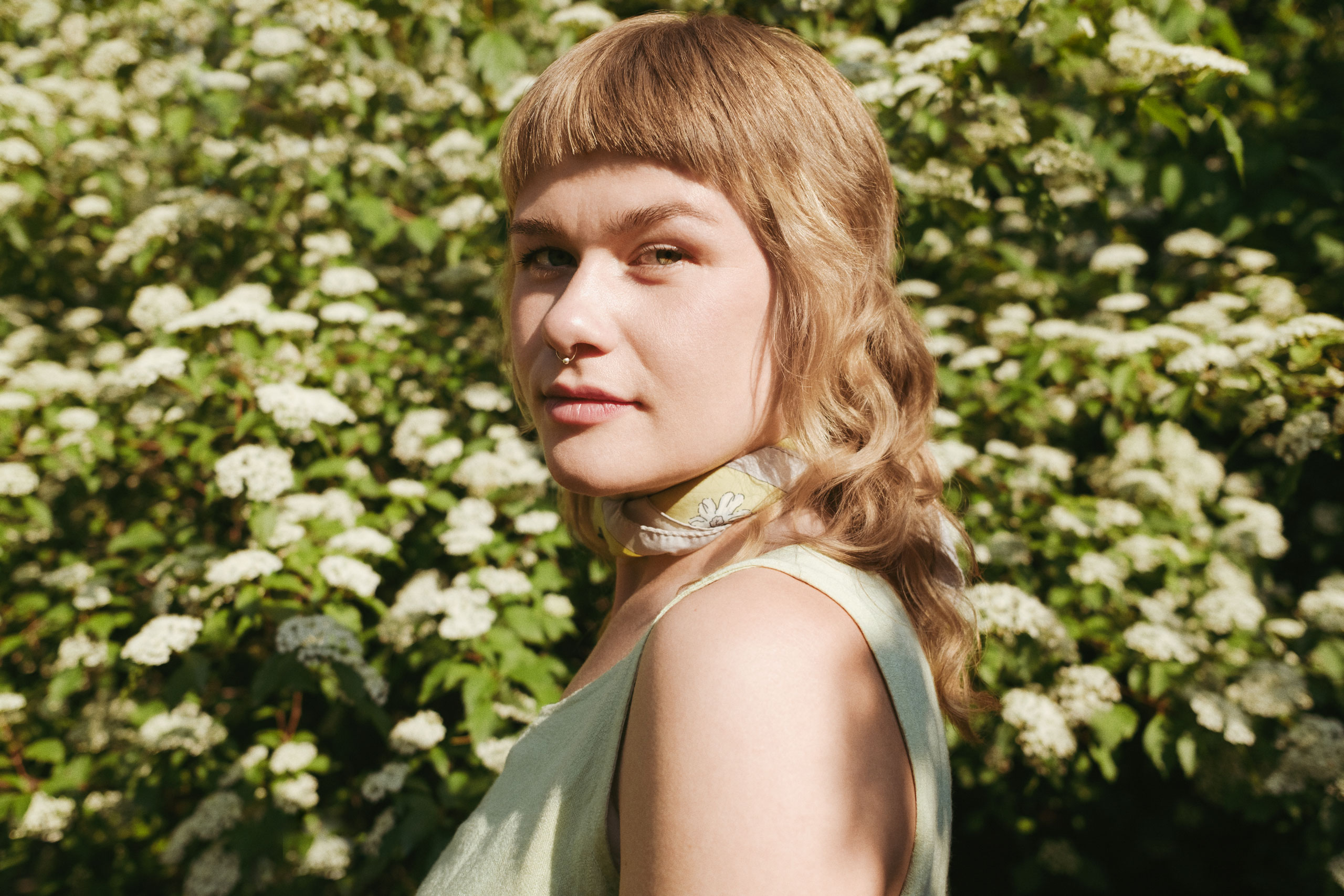 Close-up portrait of a young woman standing outdoors in front of a dense backdrop of white blooming flowers and green foliage. She has light brown wavy hair with short bangs, wears a soft yellow sleeveless top and a floral-patterned scarf around her neck. A septum piercing adds a modern touch to her natural, serene expression.