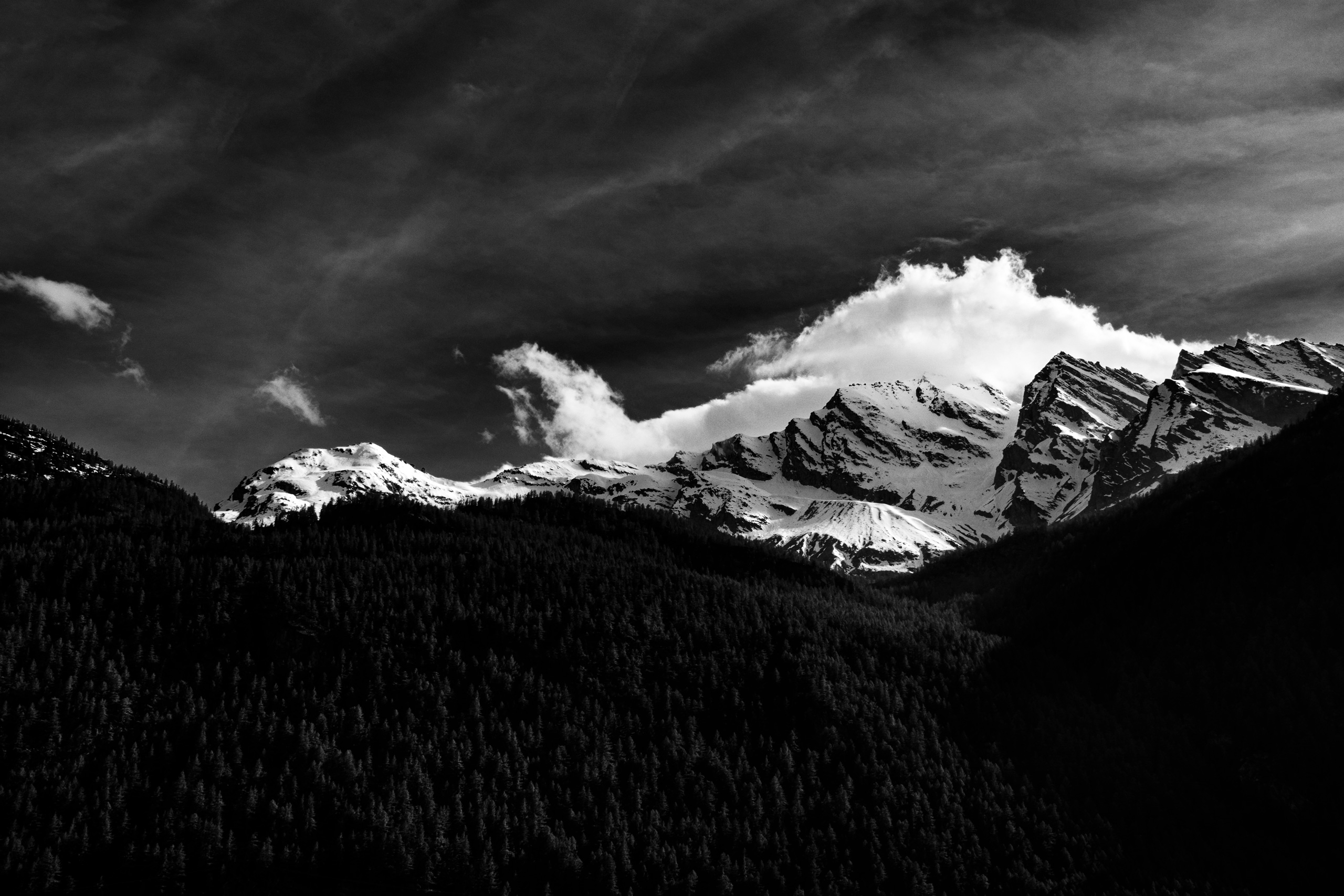 Dramatic black and white landscape photograph of a snow-covered mountain range with rugged peaks partially illuminated by sunlight, casting deep shadows across the densely forested valley below. A single cloud drapes the highest summit, adding contrast and atmosphere against the textured sky.