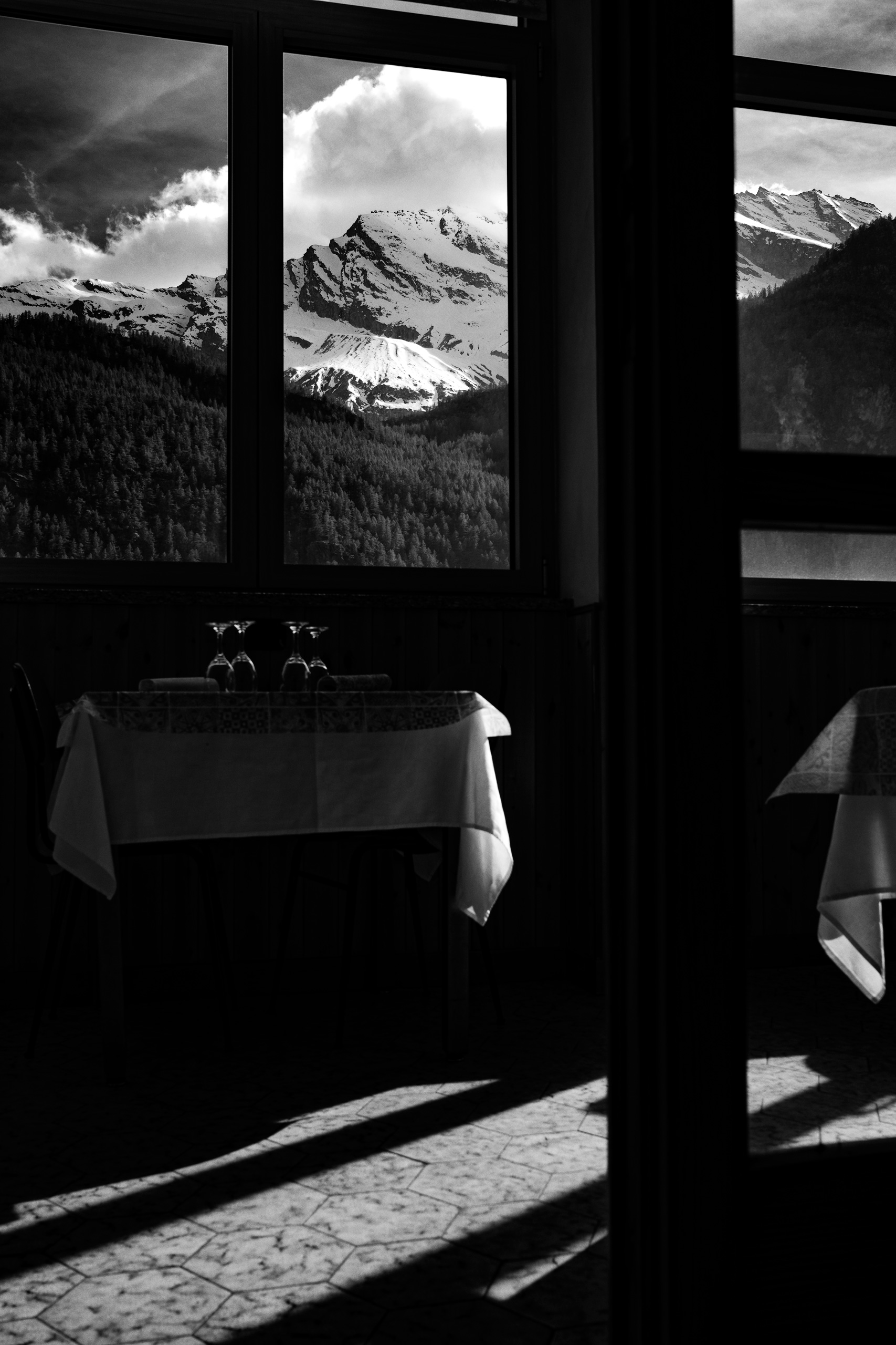 Black and white interior photograph of a dining room with a table set for two, featuring wine glasses and a tablecloth, dramatically lit by natural sunlight casting long shadows across the tiled floor. Outside the large window, a stunning view of snow-capped alpine mountains and a dense pine forest is framed, contrasting the intimate indoor ambiance with the vast, rugged outdoor landscape.