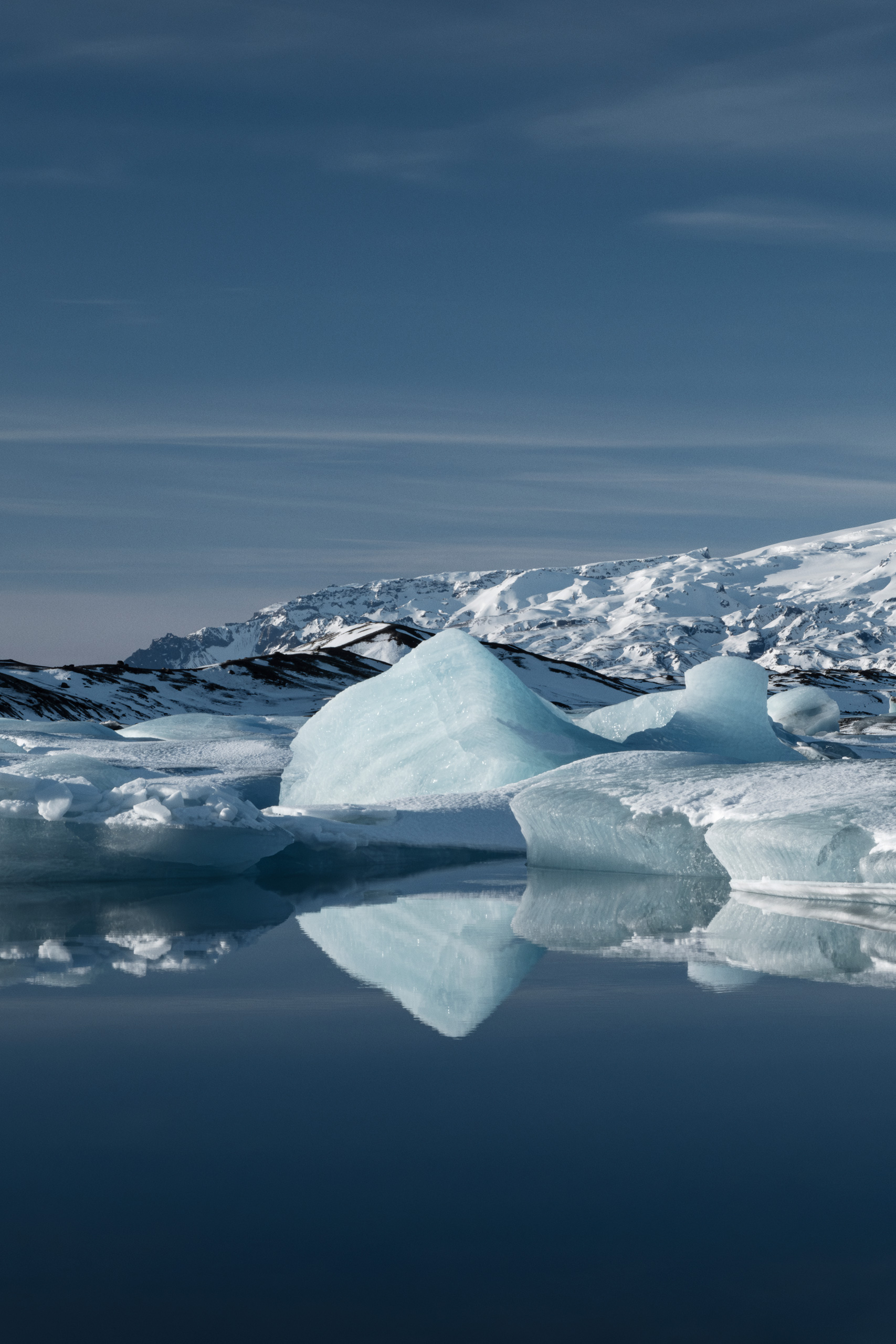 Vertical photograph of a serene arctic landscape featuring large blue and white icebergs floating on a glass-like glacial lagoon with crisp reflections. Snow-covered mountains rise in the background beneath a deep blue sky with wispy clouds.