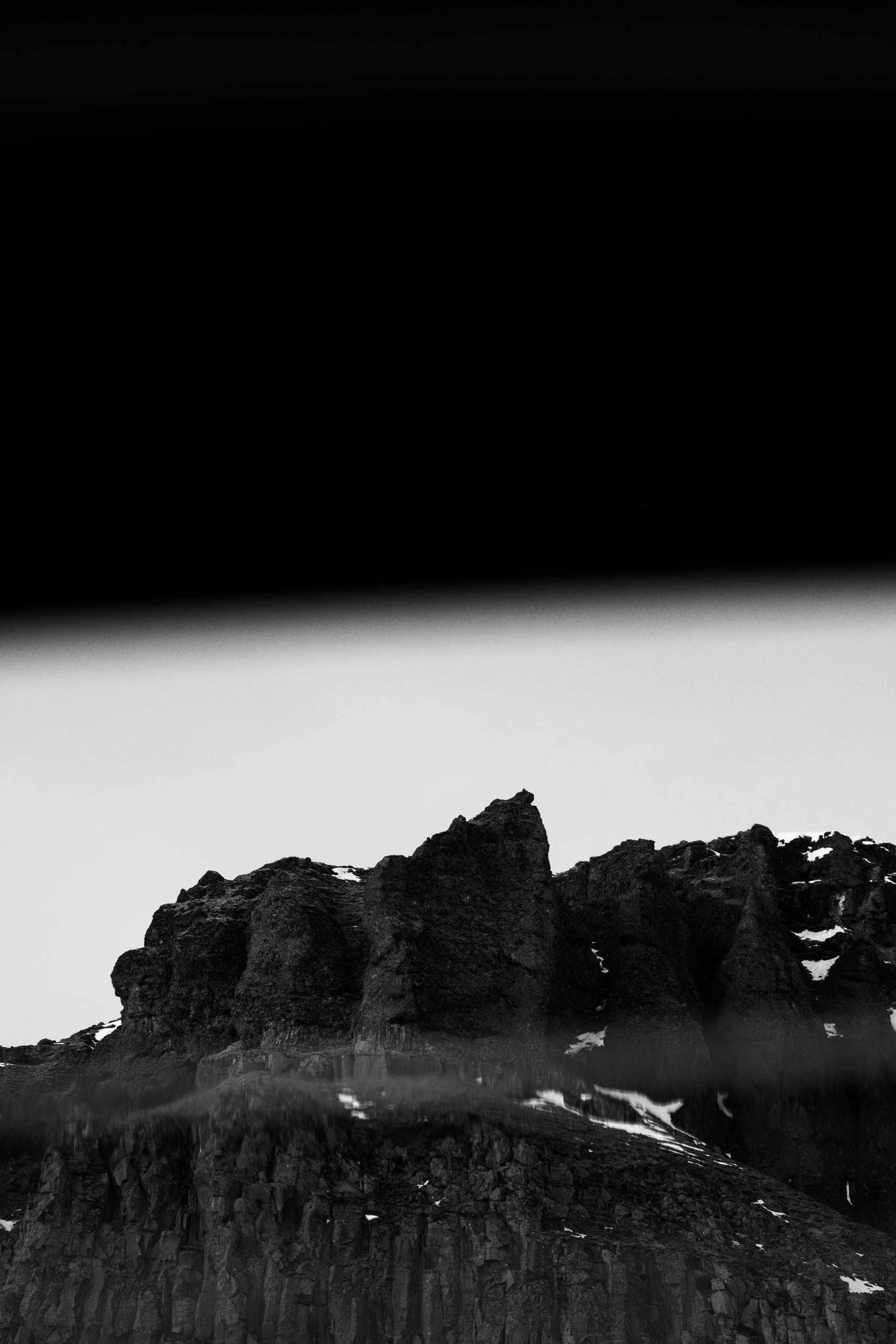 Black and white photograph of rugged volcanic rock formations with vertical cliff faces, lightly dusted with snow, reflected in a blurred water surface at the bottom of the image. The top half is a deep black gradient fading into pale sky, emphasizing contrast and abstract minimalism.