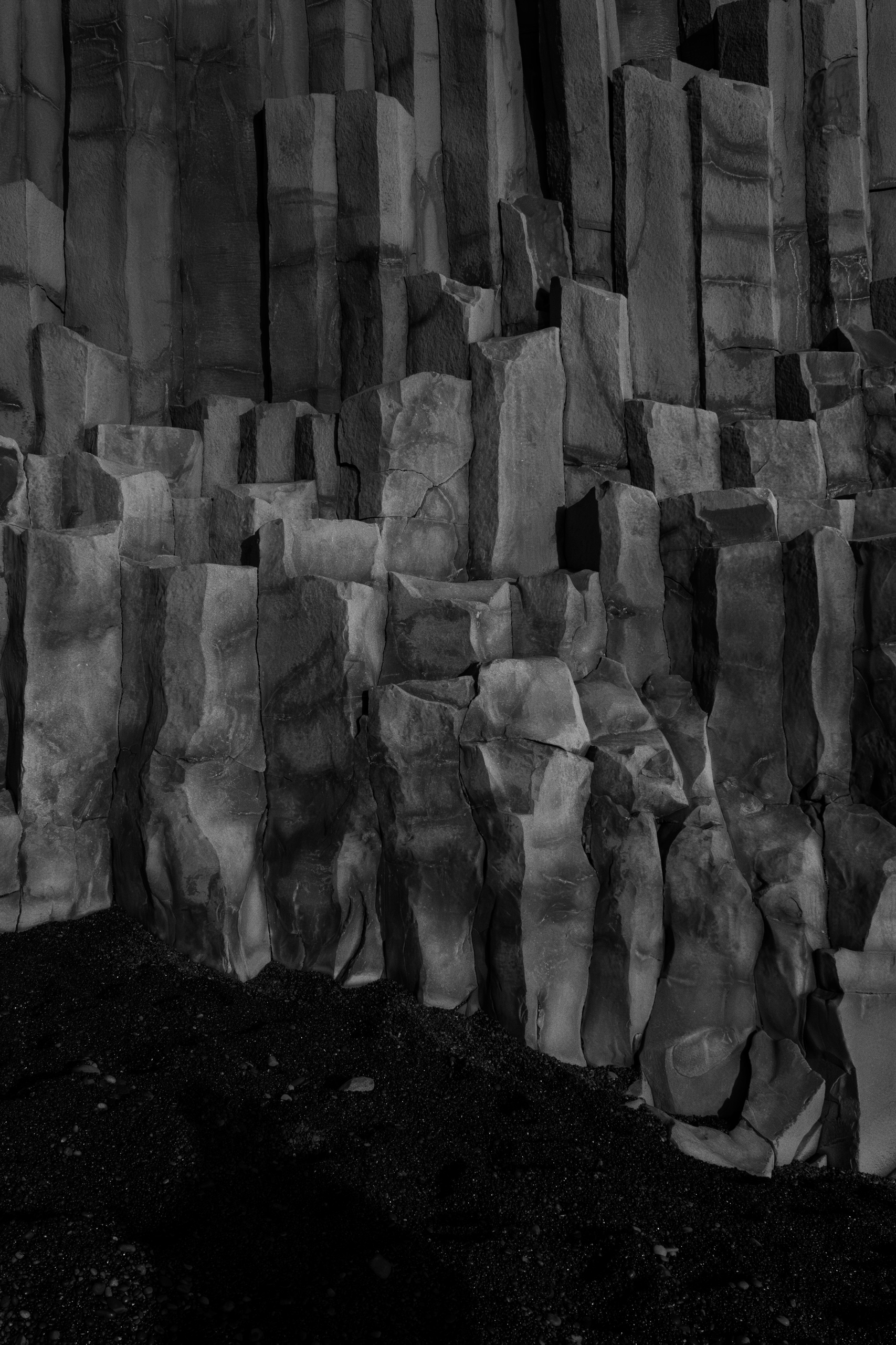 Close-up black and white photograph of naturally formed basalt columns rising vertically from a dark, pebbled ground, showcasing geometric volcanic rock formations with a striking pattern of hexagonal and rectangular shapes. The surface texture reveals erosion lines and cracks, emphasizing the geological structure.