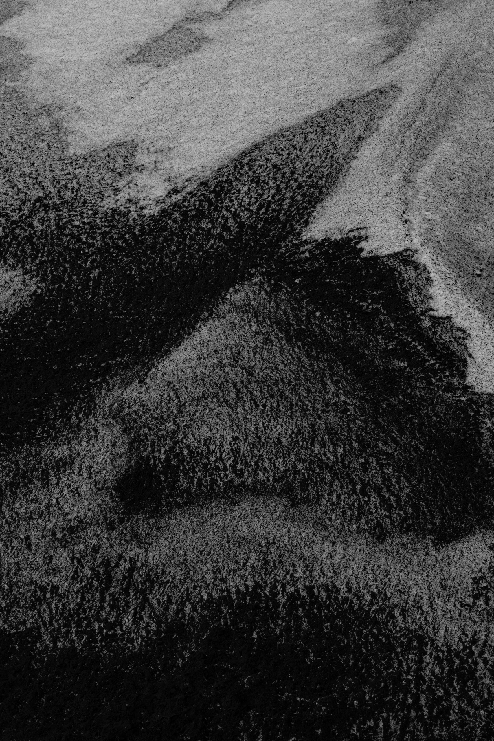 Abstract black and white close-up image of a volcanic black sand beach with fine grains creating natural patterns and textures. The photograph showcases contrasting areas of dark and light sand, shaped by wind or retreating water, forming organic lines and gradients.