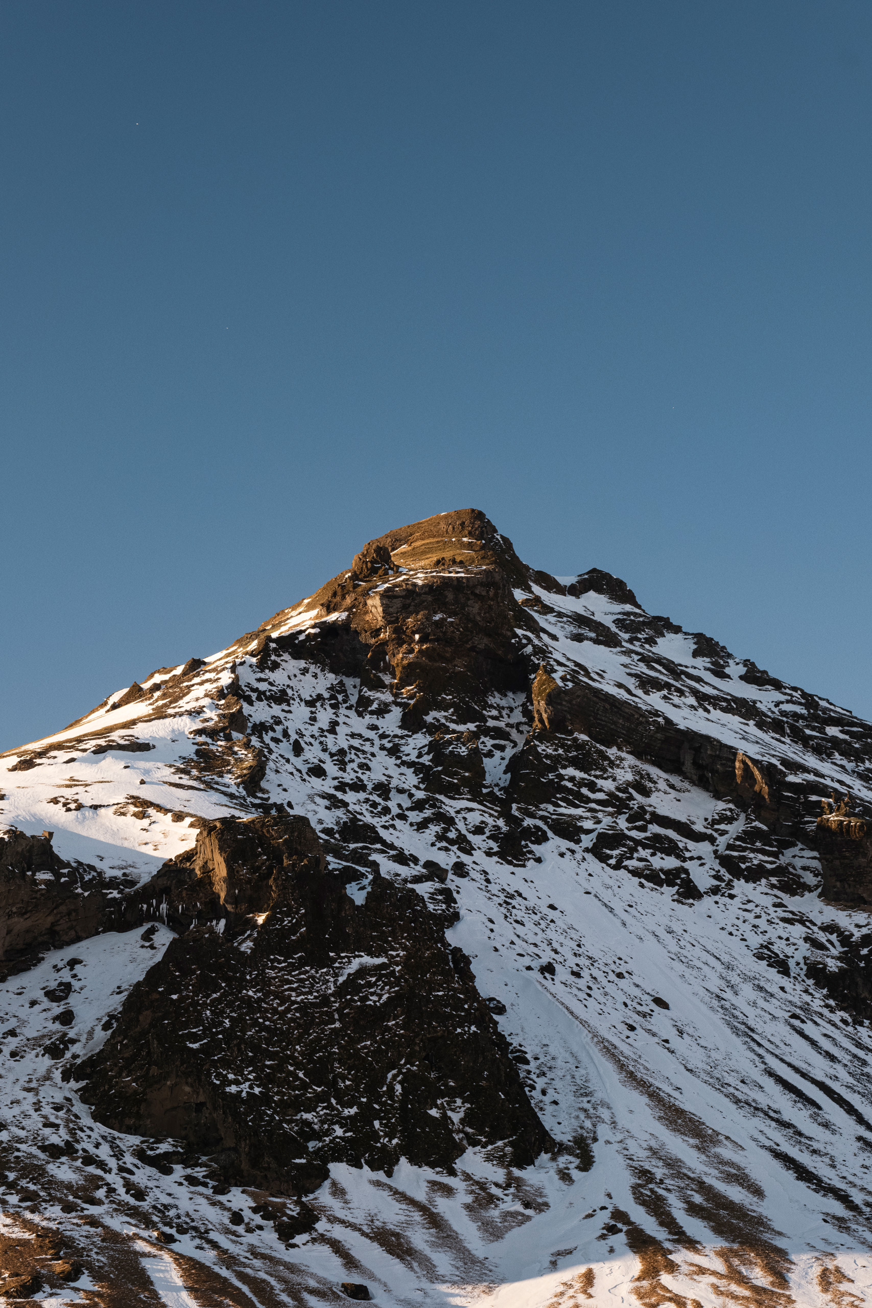 Close-up photograph of a rugged mountain peak partially covered in snow under a clear blue sky, captured during golden hour. The sunlight casts a warm glow on the rocky summit, emphasizing the textured terrain and contrasting the dark rock with the white snow patches.