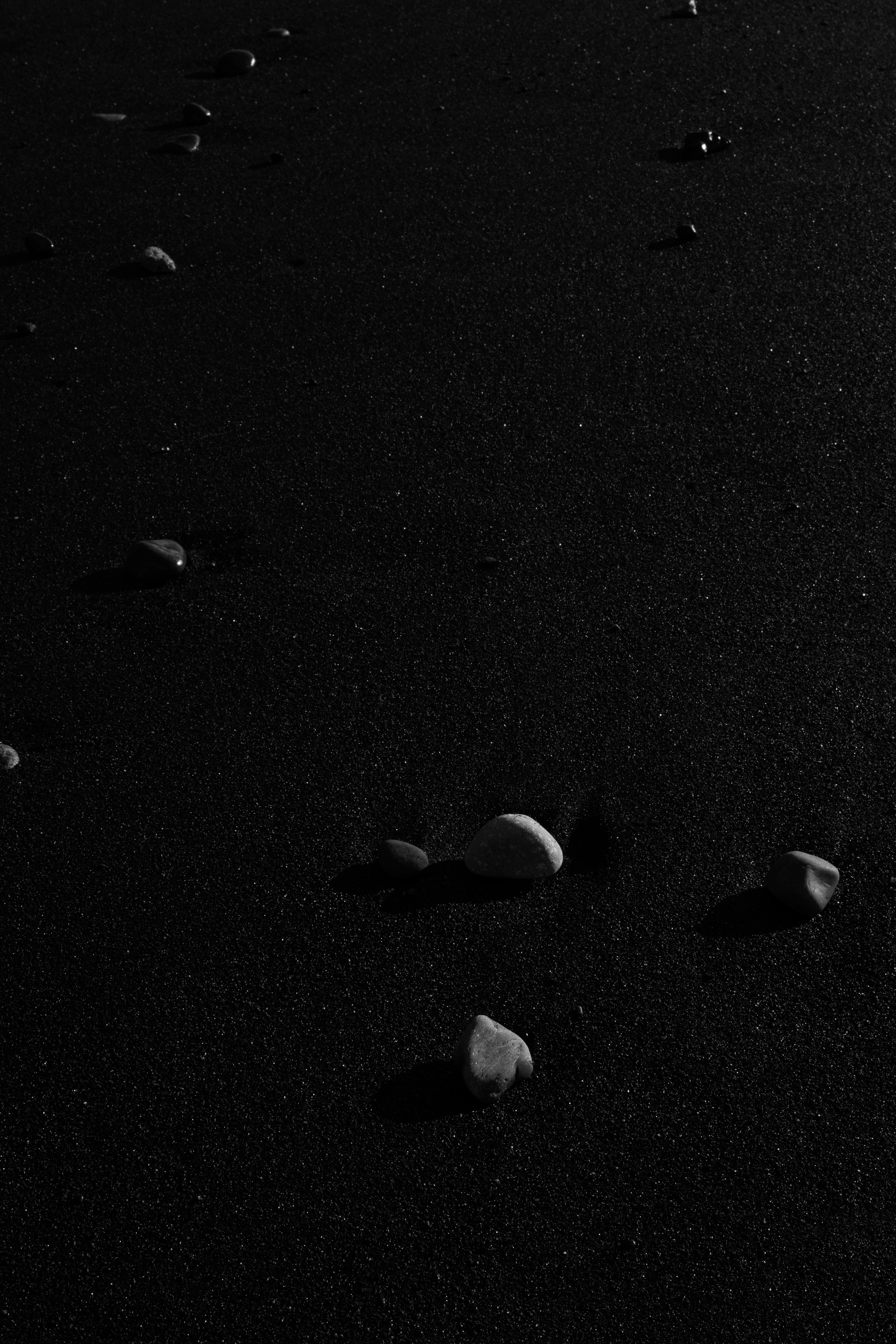 Minimalist black and white photograph of smooth stones scattered across a black sand beach, with long shadows cast by low-angle light. The granular volcanic sand surface contrasts against the rounded pebbles, creating a dramatic play of light and texture.