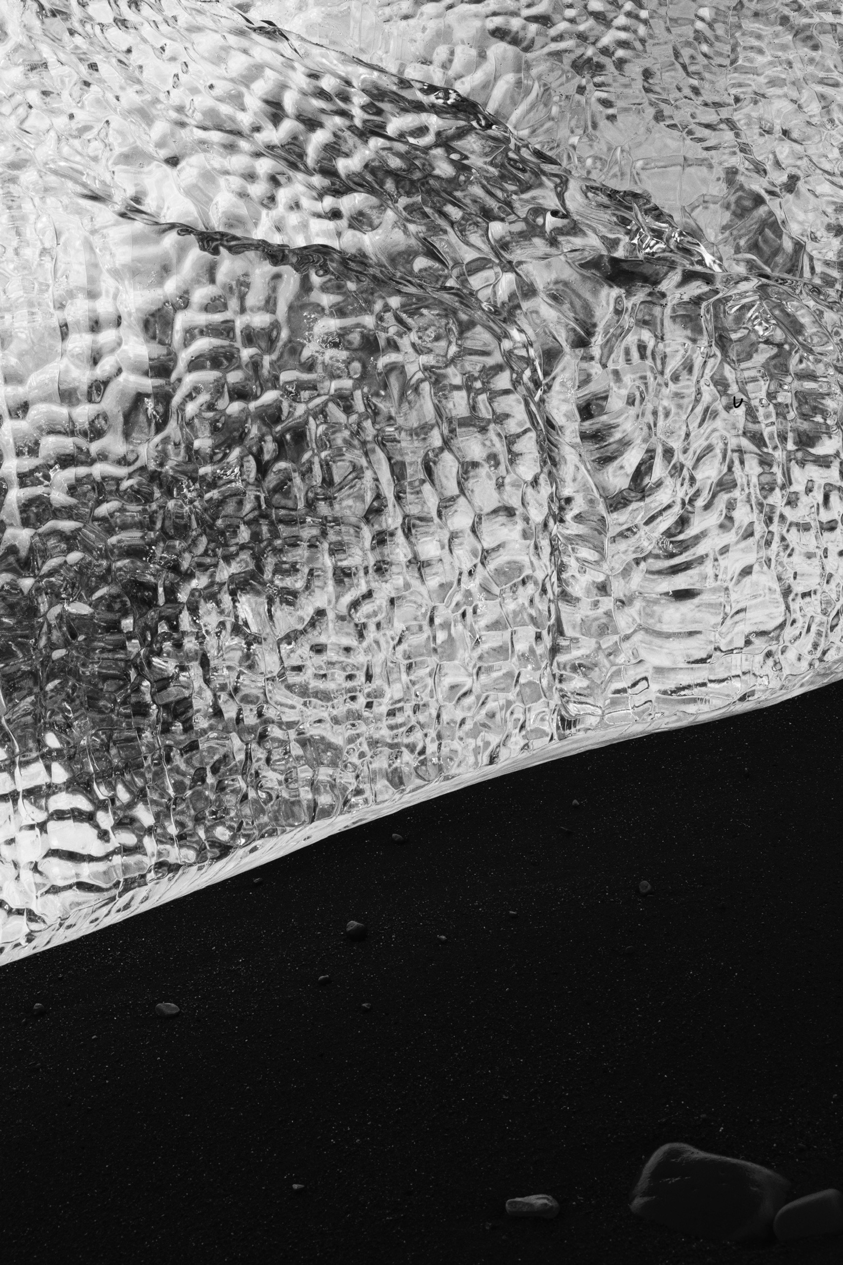 Close-up black and white photograph of a large, intricately textured piece of clear glacier ice resting on fine volcanic black sand. The ice displays a complex, crystalline pattern with ripples and refracted light, creating a high-contrast visual against the dark granular surface.
