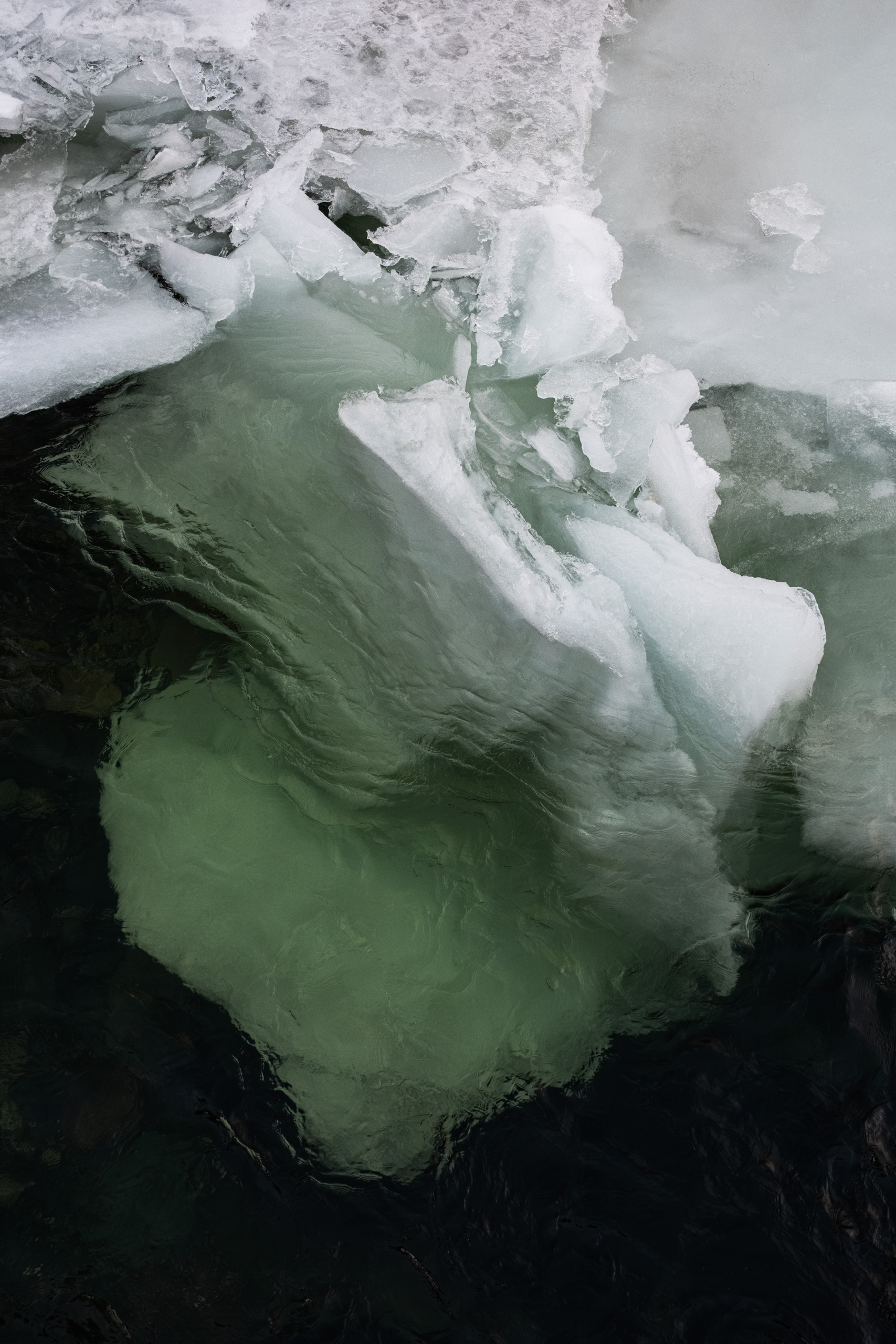 Close-up photograph of jagged, semi-submerged sea ice floating in dark Arctic waters, featuring translucent green and white hues, sharp crystalline edges, and textures formed by melting and refreezing. The layered structure reveals both surface frost and the submerged bulk of the iceberg below.