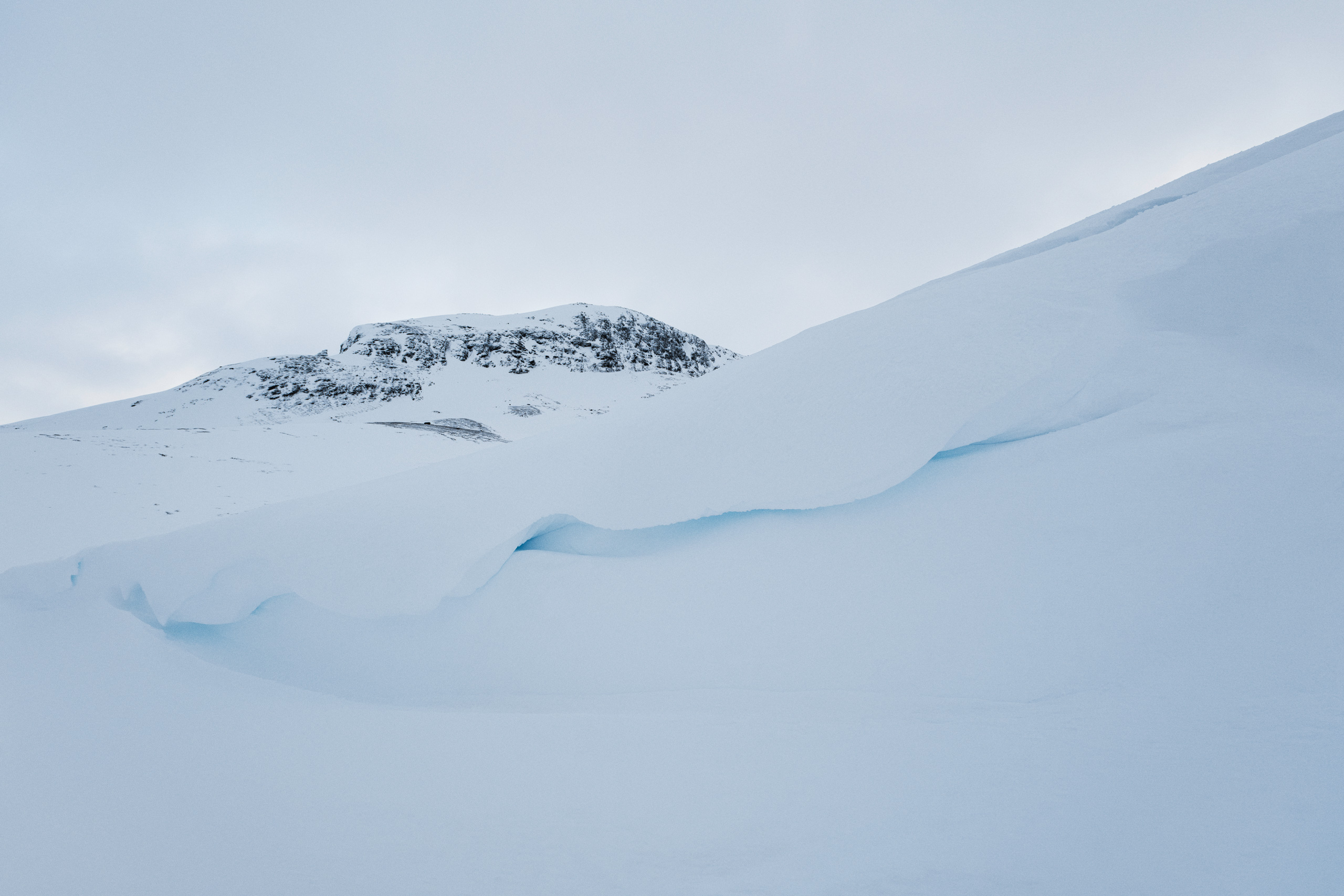 Wide-angle winter landscape featuring a snow-covered mountain with a rocky summit in the background and smooth, sculpted layers of compacted snow and glacial ice in the foreground. The icy formations create natural overhangs and ridges, with subtle blue hues indicating compressed ice beneath the snow. Overcast sky adds to the cold, arctic atmosphere.