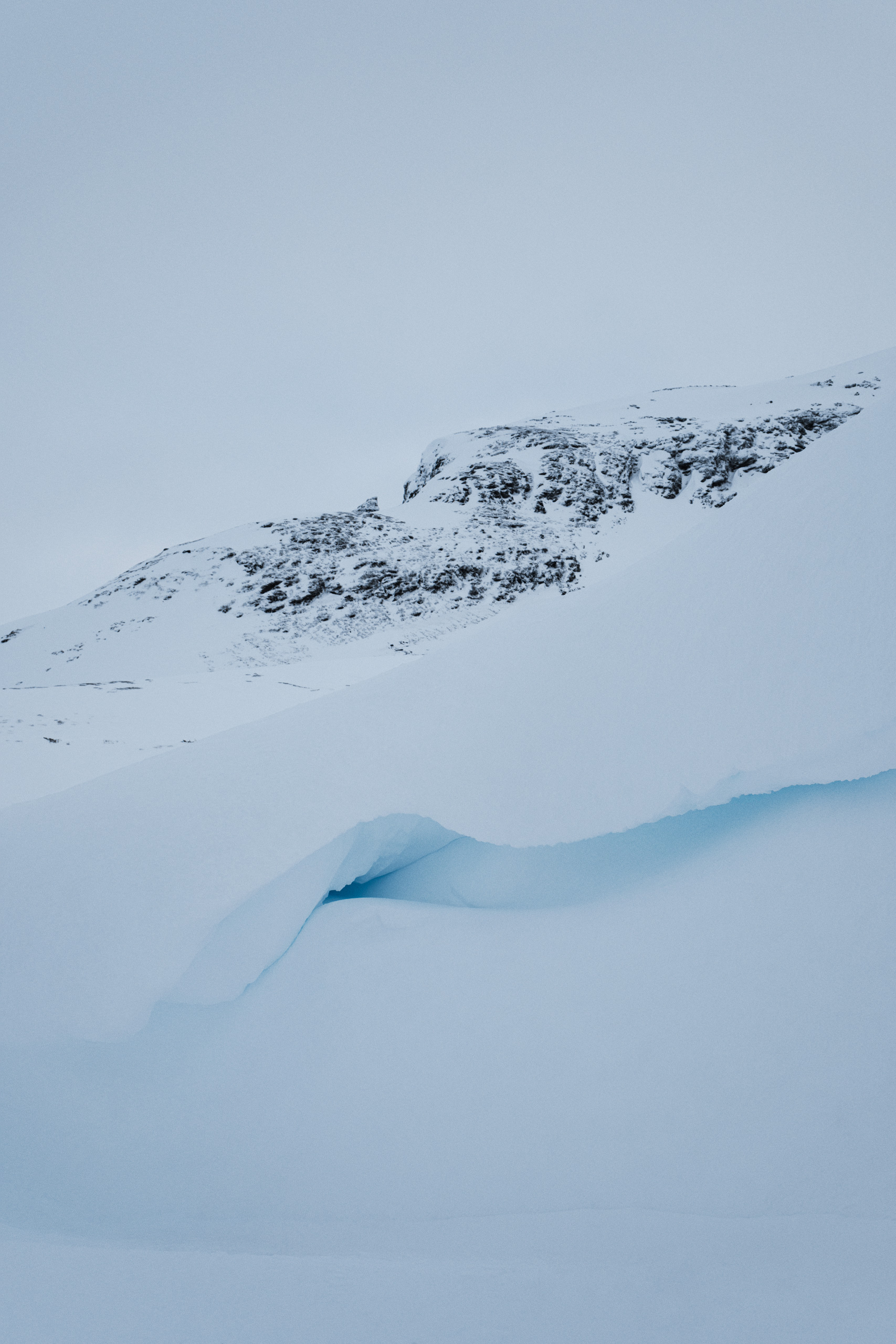 Vertical winter landscape showing a rugged, snow-covered mountain in the background and a foreground dominated by a smooth, curved snow formation with a visible crevice exposing deep blue glacial ice. The contrast between the jagged rocky slope and the soft, layered snow creates a dynamic composition under an overcast sky.