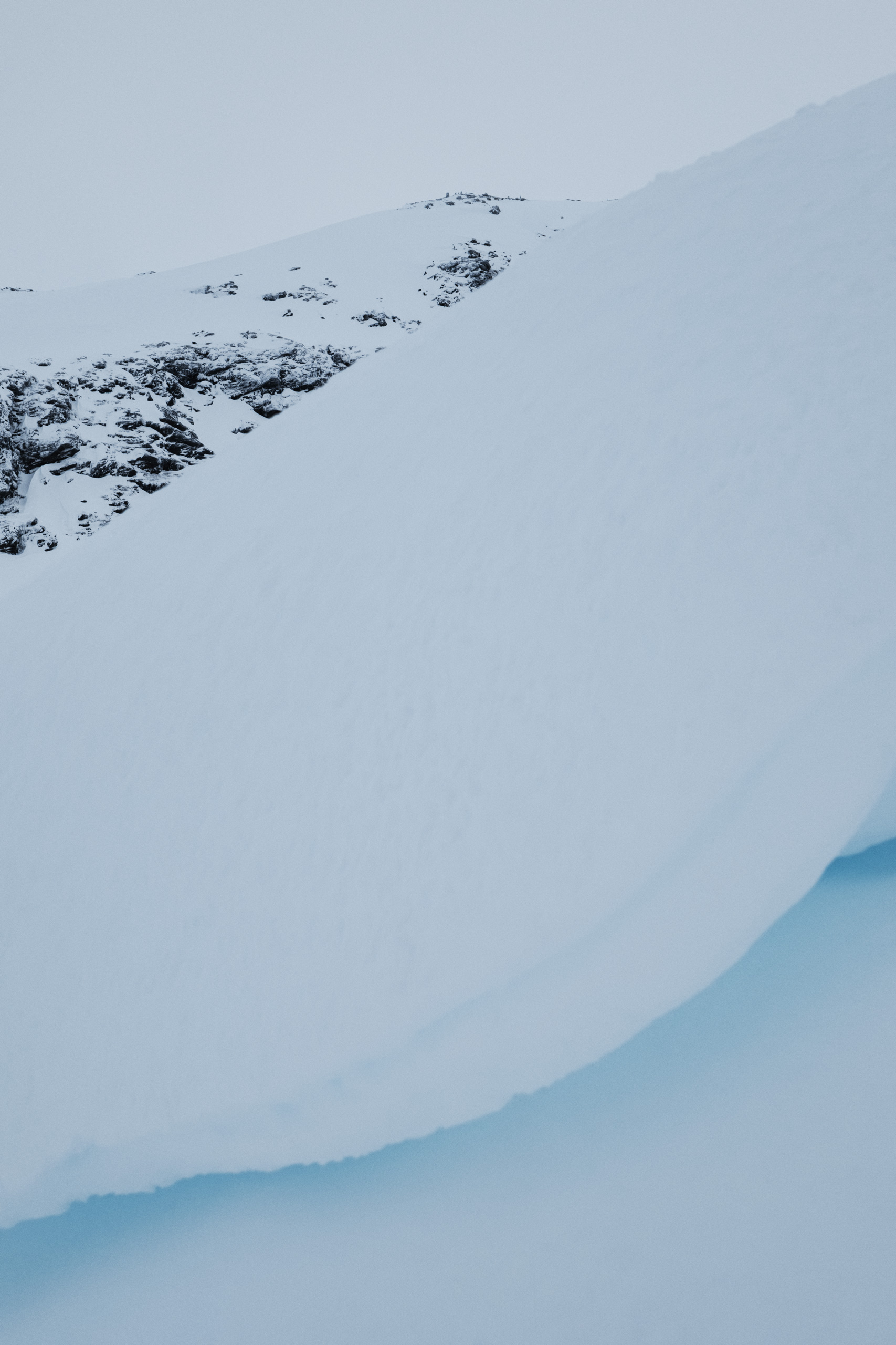Vertical photograph of a snowy mountain slope partially revealing rugged, dark rock outcrops in the upper background, contrasted by a large, smooth foreground of snow and glacial ice. A deep snowbank arcs diagonally across the frame, with subtle blue hues indicating compacted ice beneath the surface. The composition emphasizes texture, contrast, and the raw forms of a cold, alpine landscape.