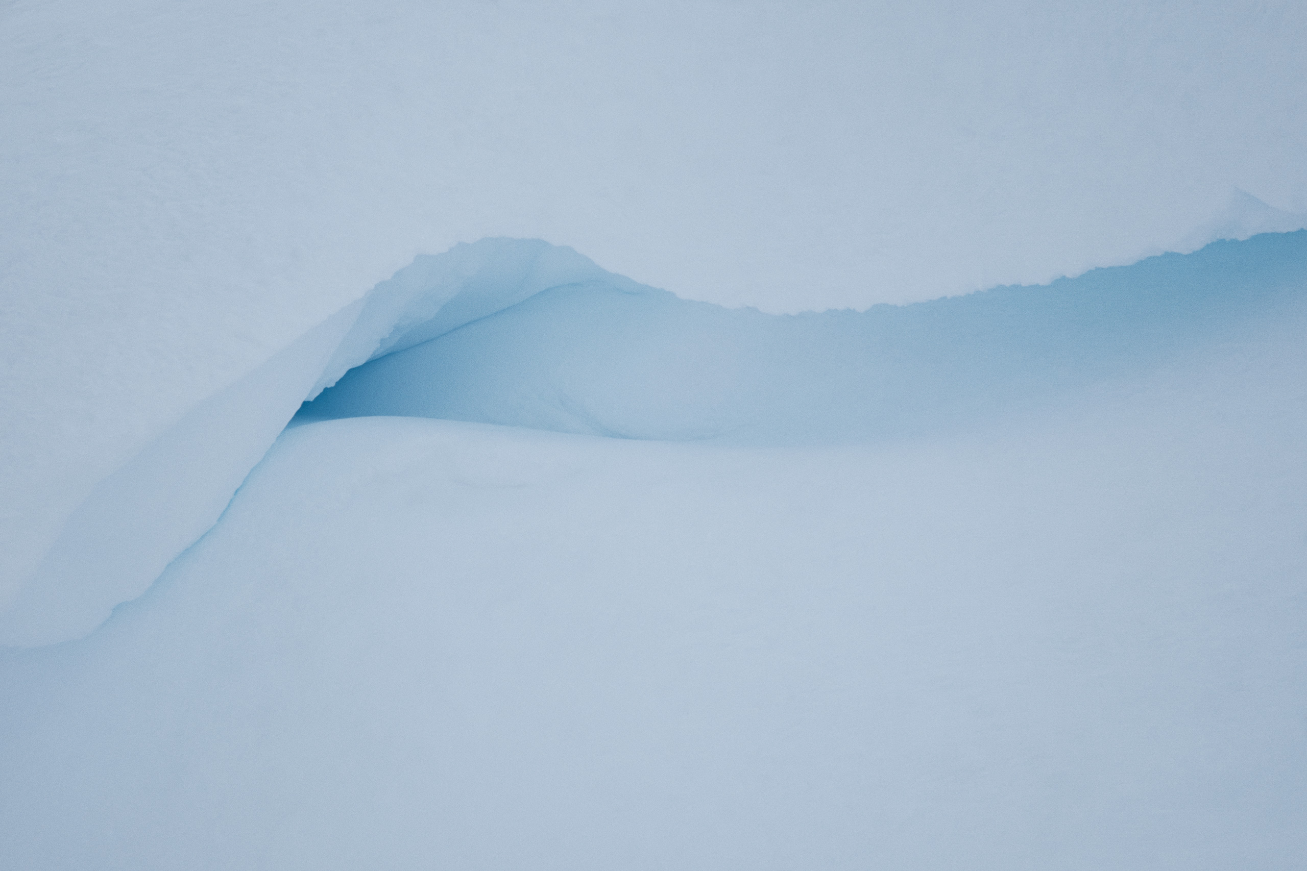Close-up photograph of a smooth snowdrift with a naturally formed, curved indentation revealing compacted blue-tinted ice beneath the surface; the soft lighting highlights the texture and subtle color gradient in the snow and ice, evoking an abstract, minimalist winter landscape.