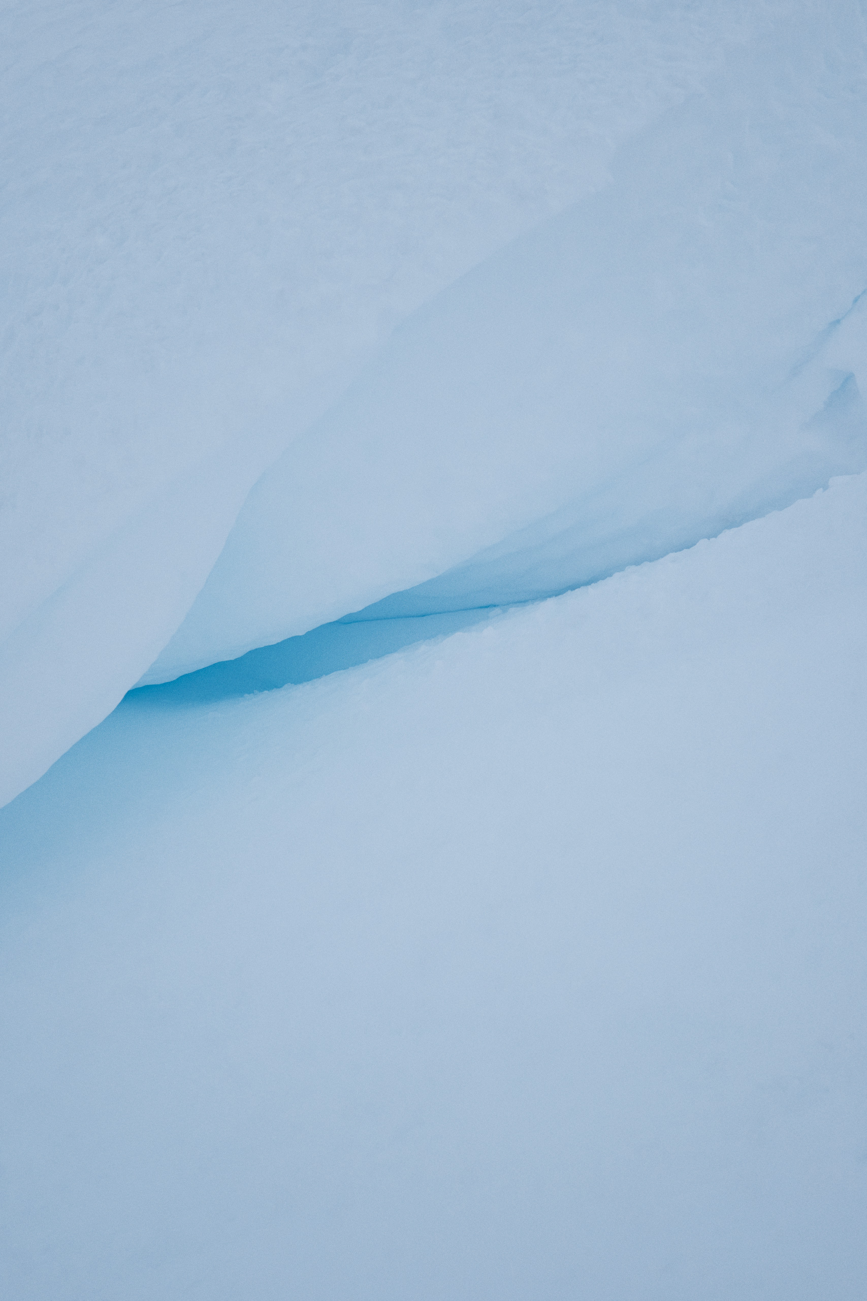 Vertical close-up of a naturally sculpted snow formation with a narrow crevice exposing dense, blue-tinted glacial ice beneath a smooth snow layer. The image highlights the subtle gradient from soft white to icy blue, emphasizing the compact texture and layered structure of the frozen landscape.